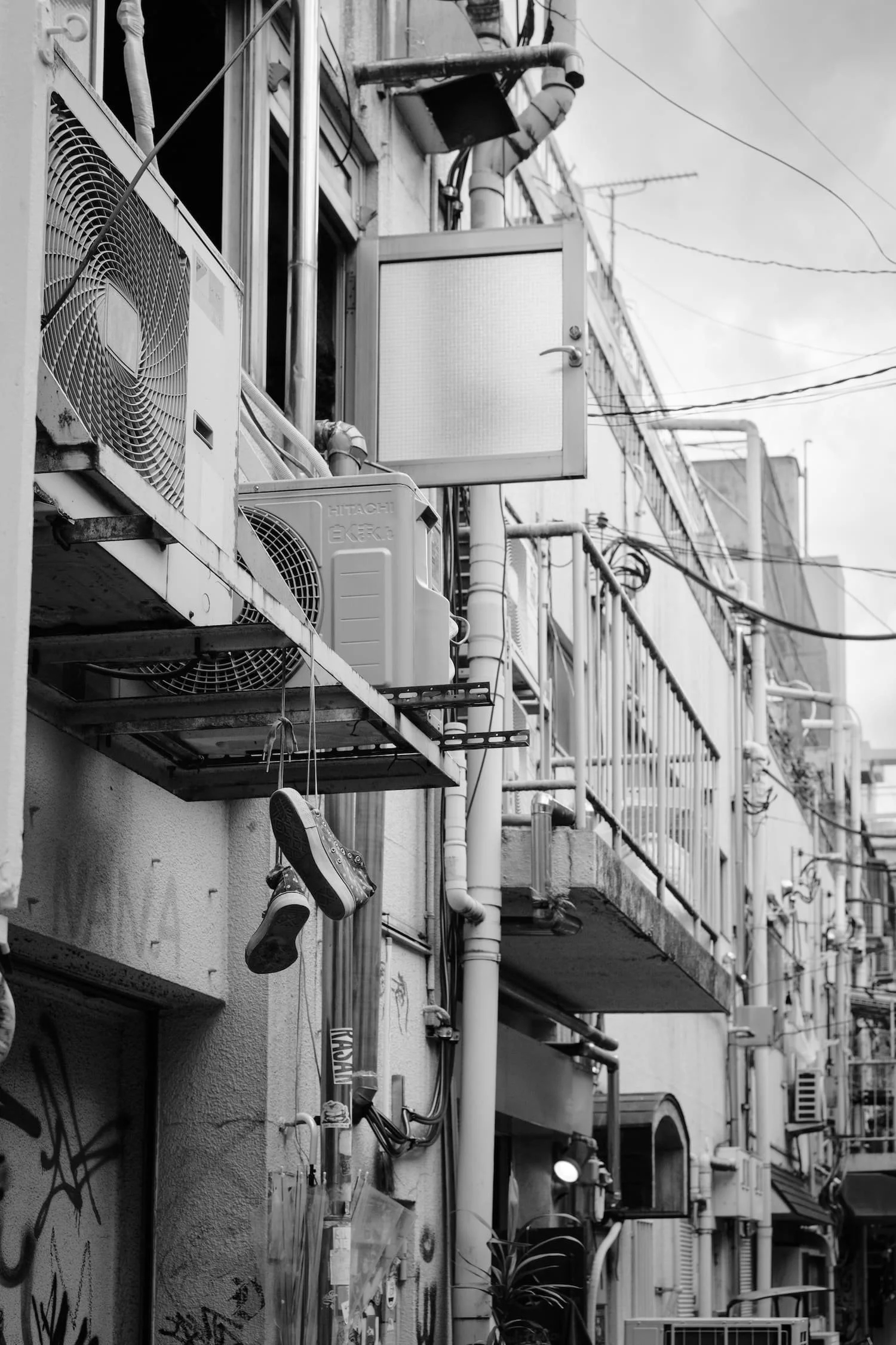 Black and white photo of an urban building exterior with air conditioning units, wires, a balcony railing, a pair of sneakers hanging from a wire, and graffiti on the wall.