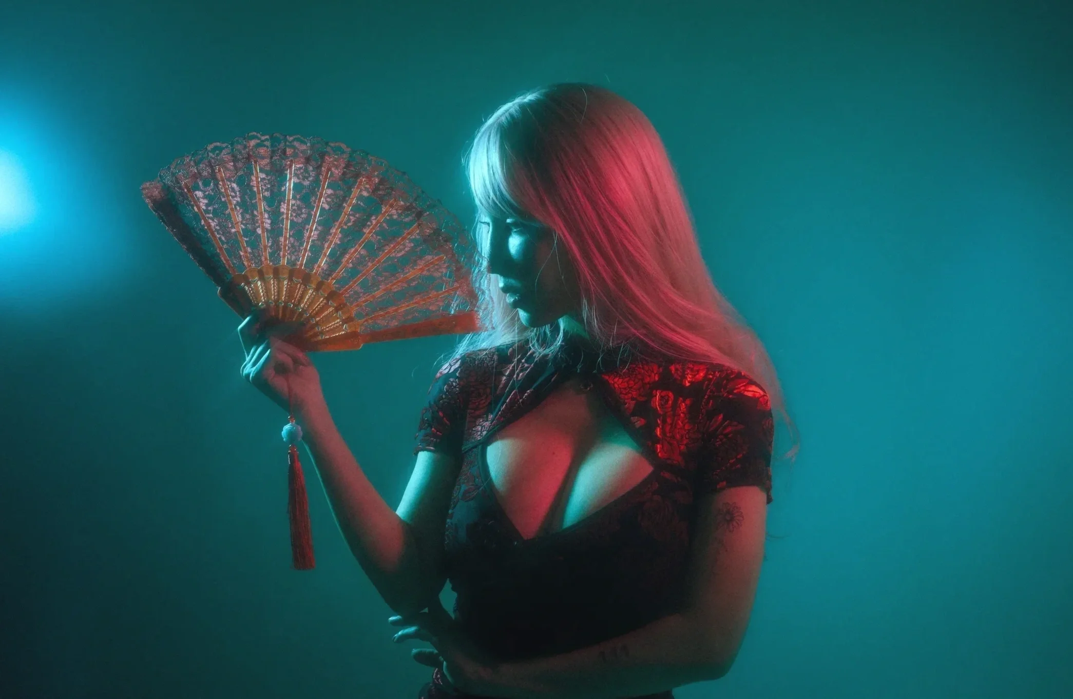 A woman with pink hair holding an ornate fan, wearing a dark dress with lace details, in a dimly lit environment with teal and pink lighting.