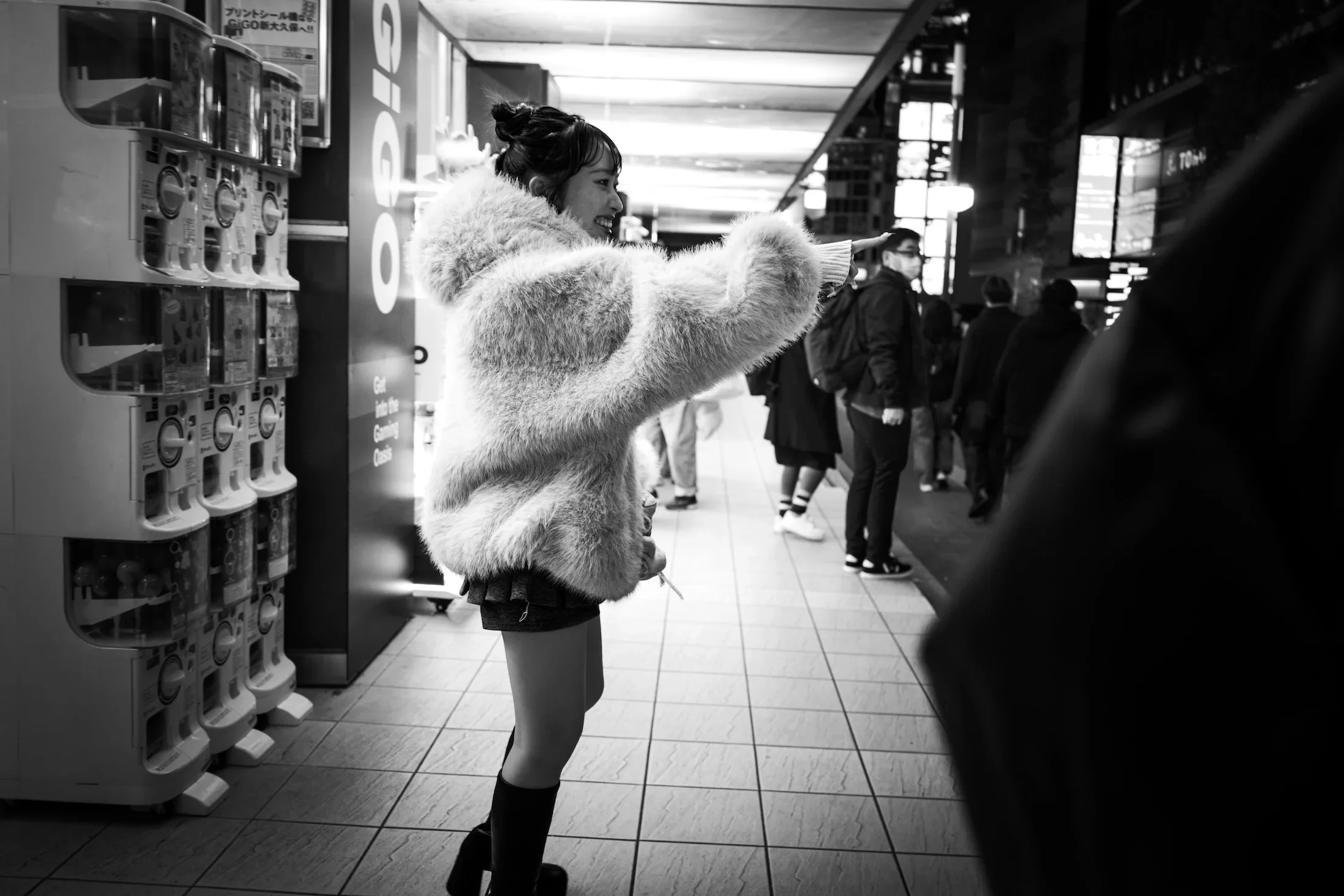 A woman in a fluffy jacket and short skirt standing at a vending machine in a train station, smiling and pointing. Other people are seen in line in the background.
