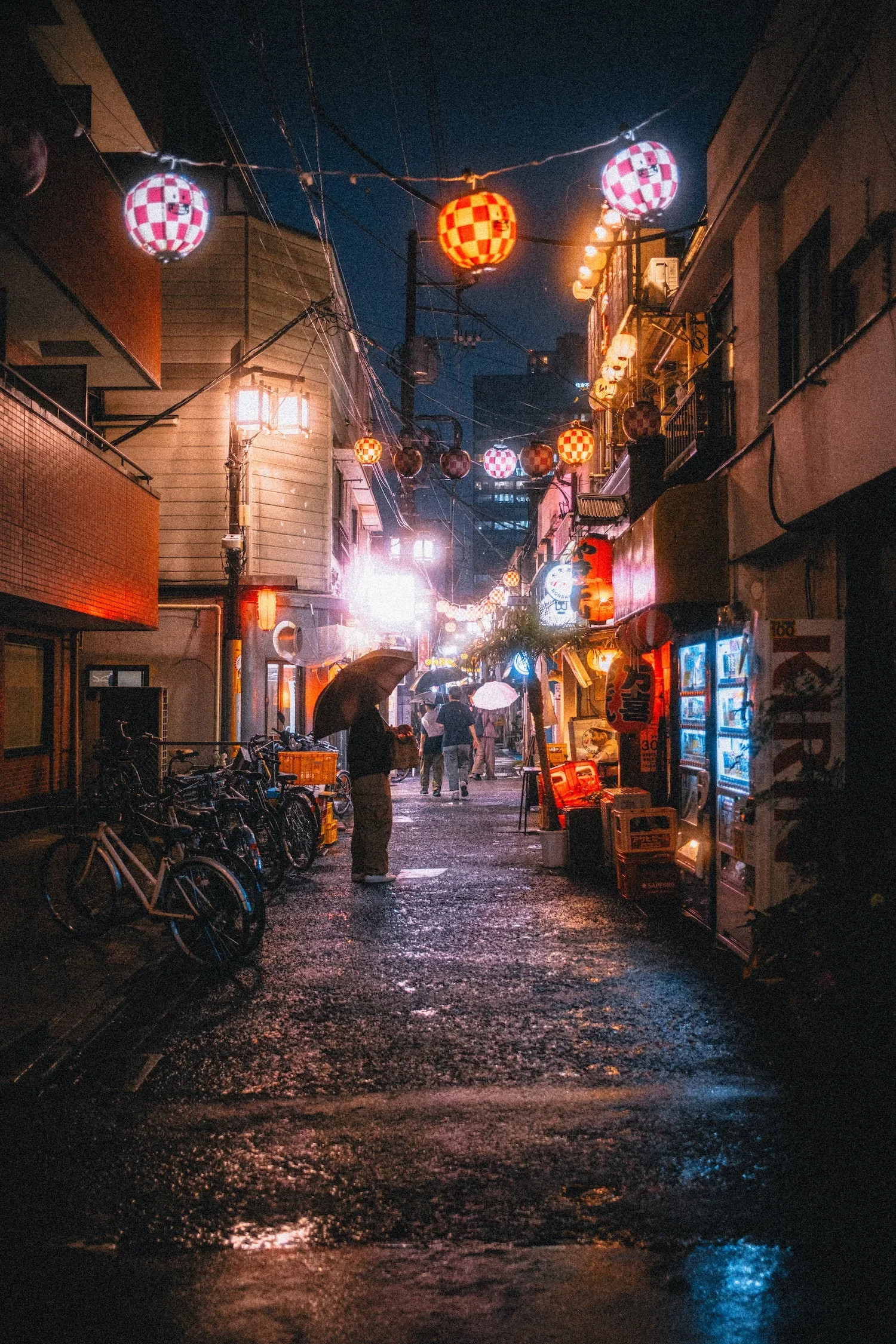 Night street scene with lanterns hanging above, people holding umbrellas walking on wet pavement, illuminated signs and storefronts, some bicycles parked on the side.