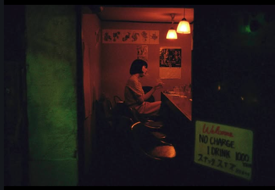 A person with short hair sitting alone at a bar counter, reading a menu in a dimly lit room with hanging pendant lights, posters on the wall, and a sign that says 'No Charge 1 Drink 1000' in the foreground.