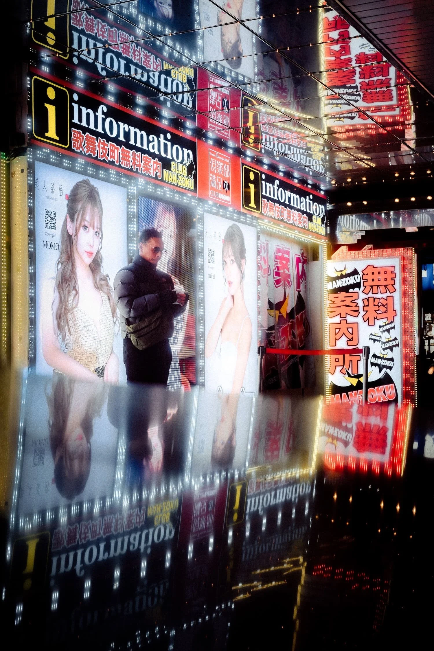 Brightly lit advertisements and signs with images of women and a man, featuring Japanese and English text, in an urban night setting with reflections on the ground.