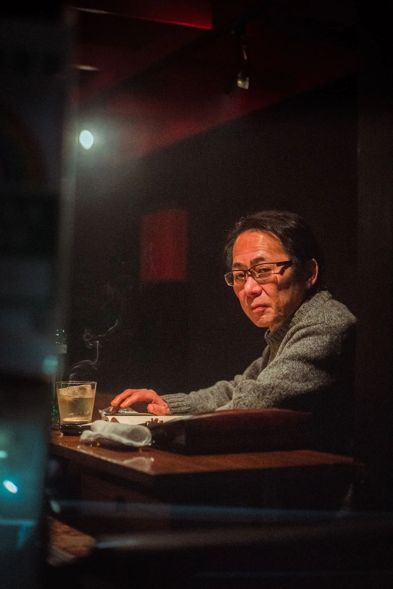 A man with glasses and gray sweater sitting at a wooden table in a dimly lit restaurant, with a glass of steaming beverage near his hand and a closed laptop in front of him.