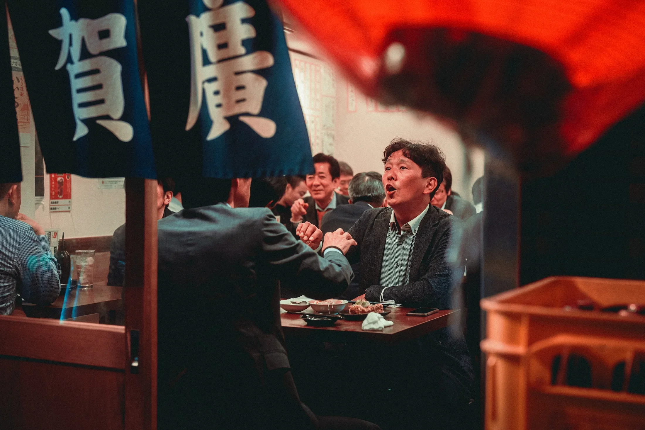 A group of people are sitting and talking at a restaurant, with blue Japanese banners hanging in the foreground and red lighting illuminating part of the scene.