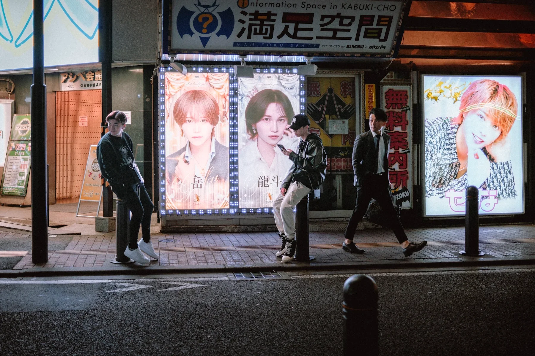 Night scene of a city street with four young men waiting at a bus stop in front of bright illuminated posters and signs. The posters feature images of a male and female celebrities, with the female having short light-colored hair. The environment appears urban with storefront signs and advertisements in the background.