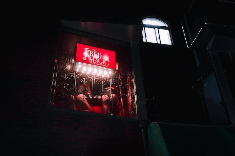 A window display titled 'The Red Magic' with a person shown from the waist down, sitting on a red velvet couch behind a decorative metal balcony, illuminated by bright lights, at night.