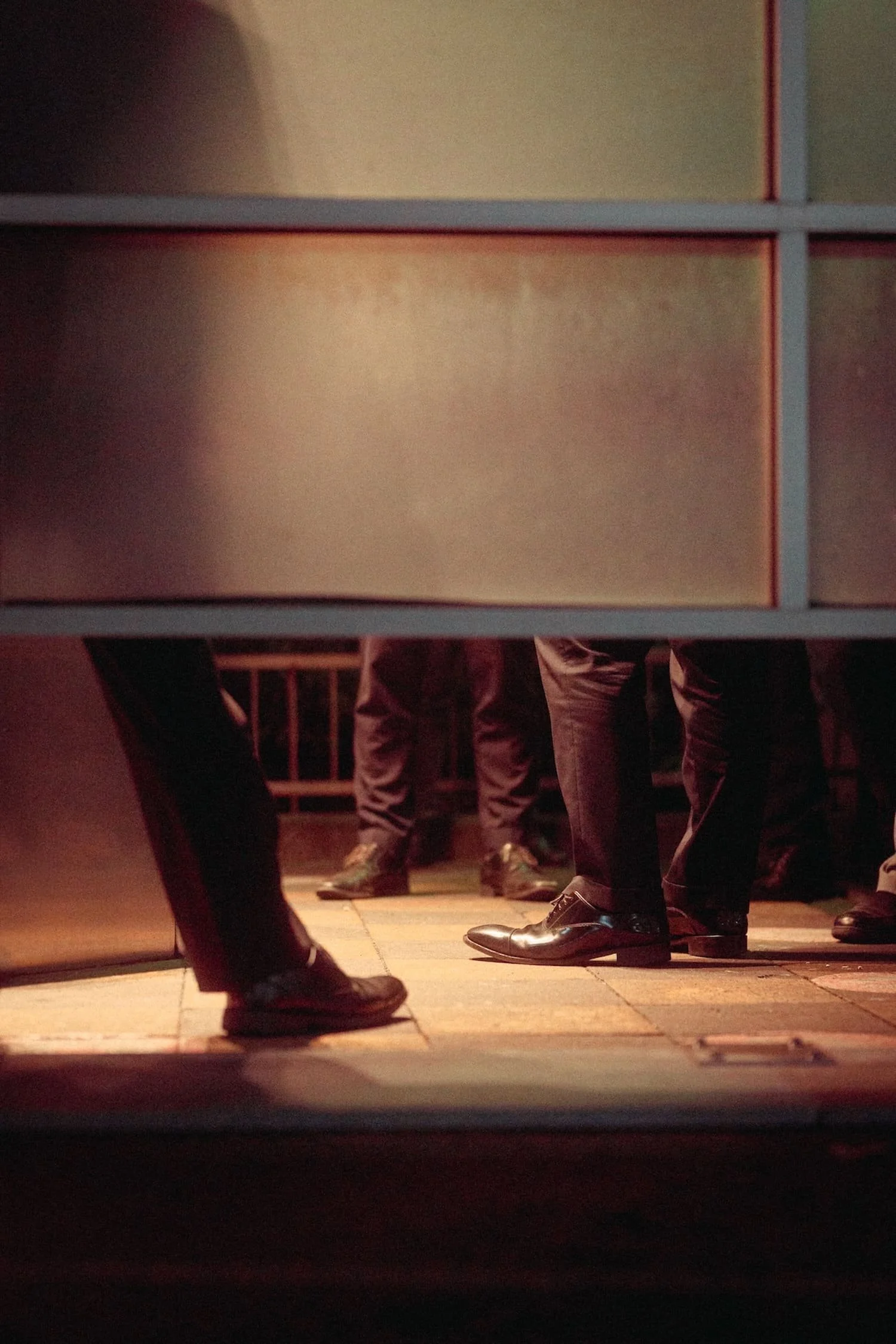 People in formal attire, wearing dark suits and shoes, are gathered in a dimly lit room, viewed from a low angle beneath a table or partition.