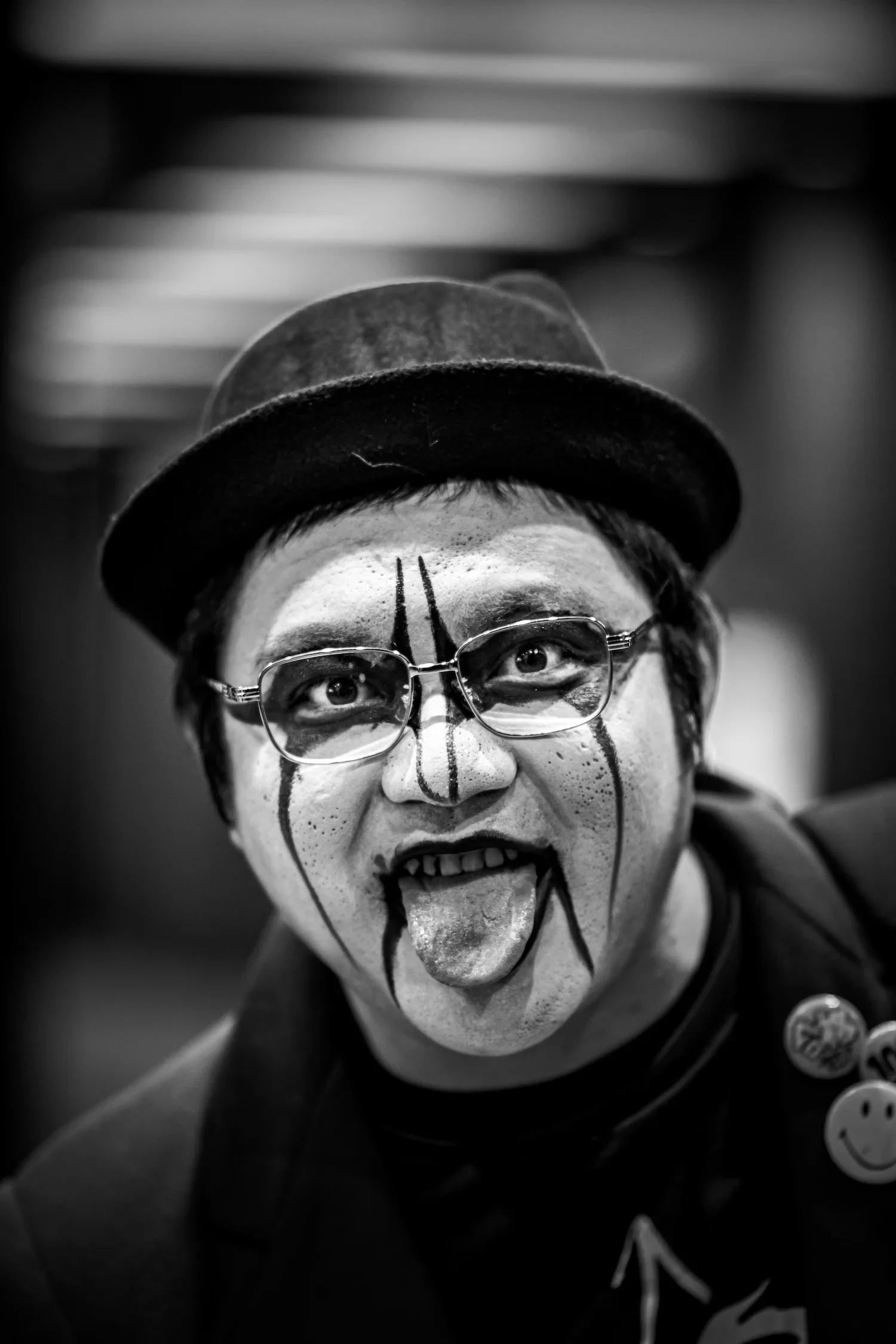 A person with clown makeup is sticking out their tongue and making a goofy face, wearing glasses, a hat, and a dark shirt with a smiley face badge.