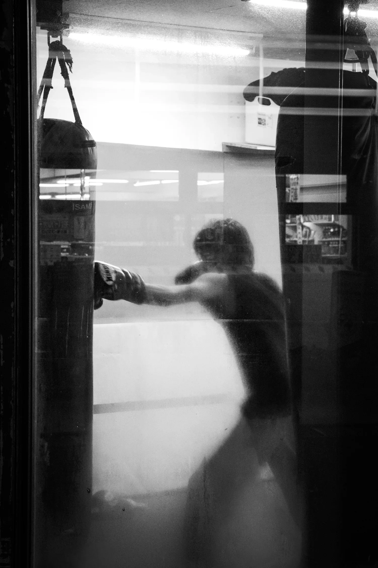 A person throwing a punch at a punching bag in a gym, reflected in a window with a darkened room and a car outside visible.