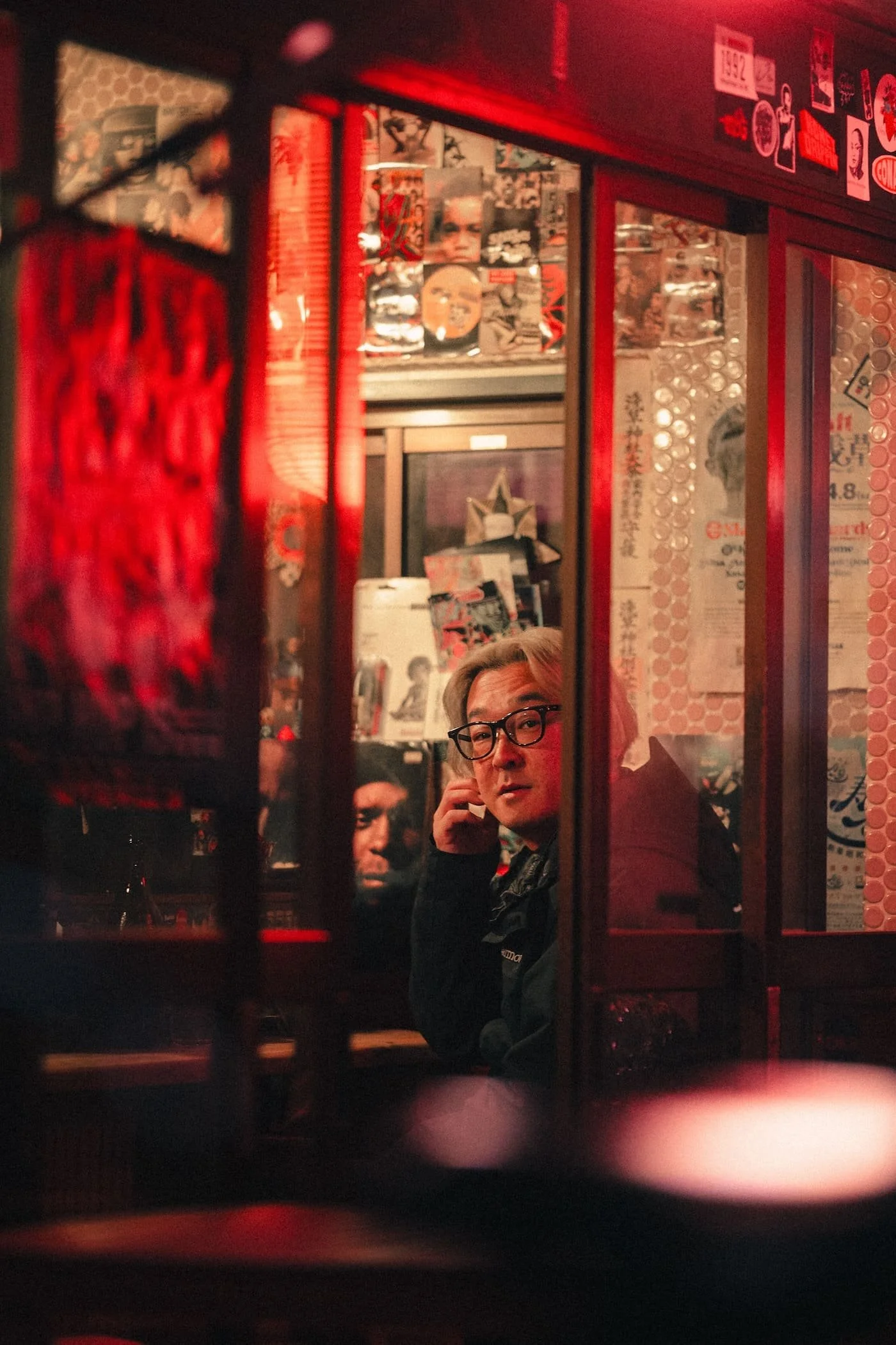 A person with glasses sitting inside a colorful, decorated Asian-themed restaurant or shop, looking toward the camera through a glass window, surrounded by posters and stickers on the walls.