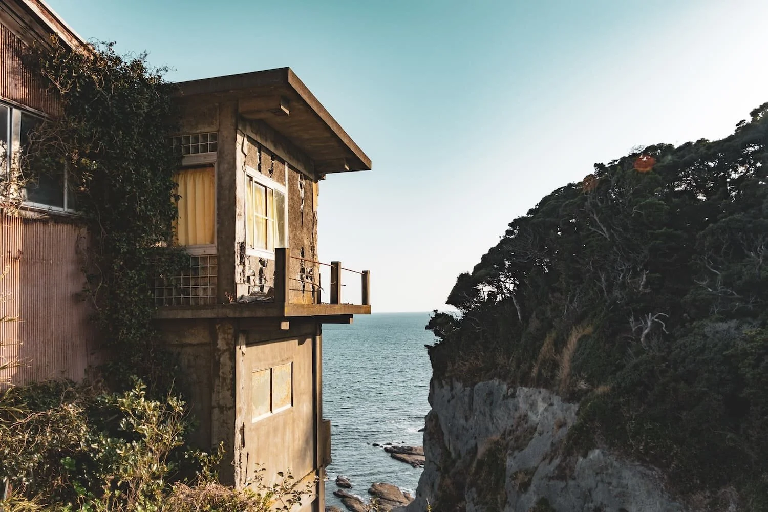 A weathered, concrete building perched on the edge of a cliff overlooking the ocean, with greenery covering part of its facade and a rocky shoreline below.