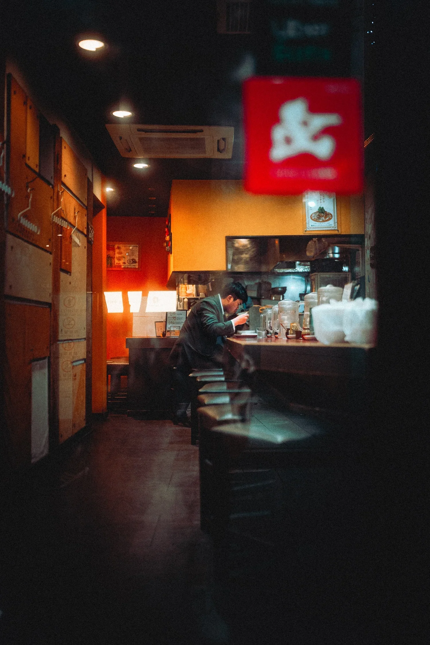 Inside a dimly lit Japanese restaurant with a man sitting alone at the counter eating, surrounded by bowls, cups, and condiments. Wooden decor and menu boards are visible in the background.
