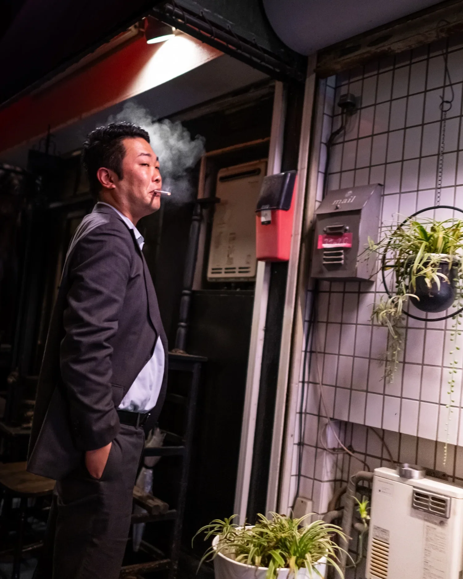 A man wearing a suit with his hands in his pockets, smoking a cigarette in an indoor space. There is a potted plant in the foreground and a tiled wall with a mailbox and electronic equipment behind him. The lighting is dim with a focused spotlight overhead.