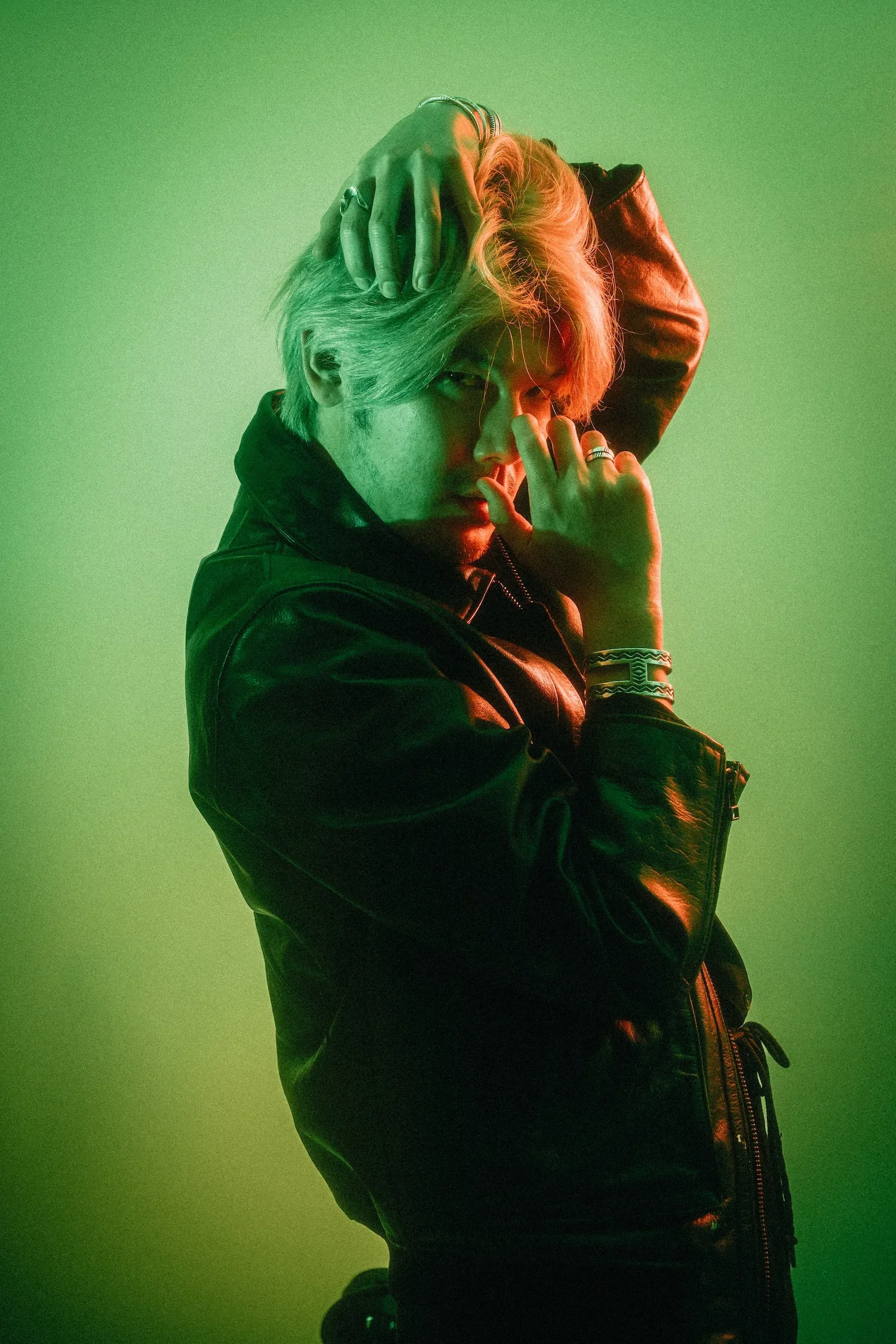 A person with blonde hair styled in loose waves, wearing multiple rings and a bracelet, in a black leather jacket, striking a pose with hands near their face against a green background.