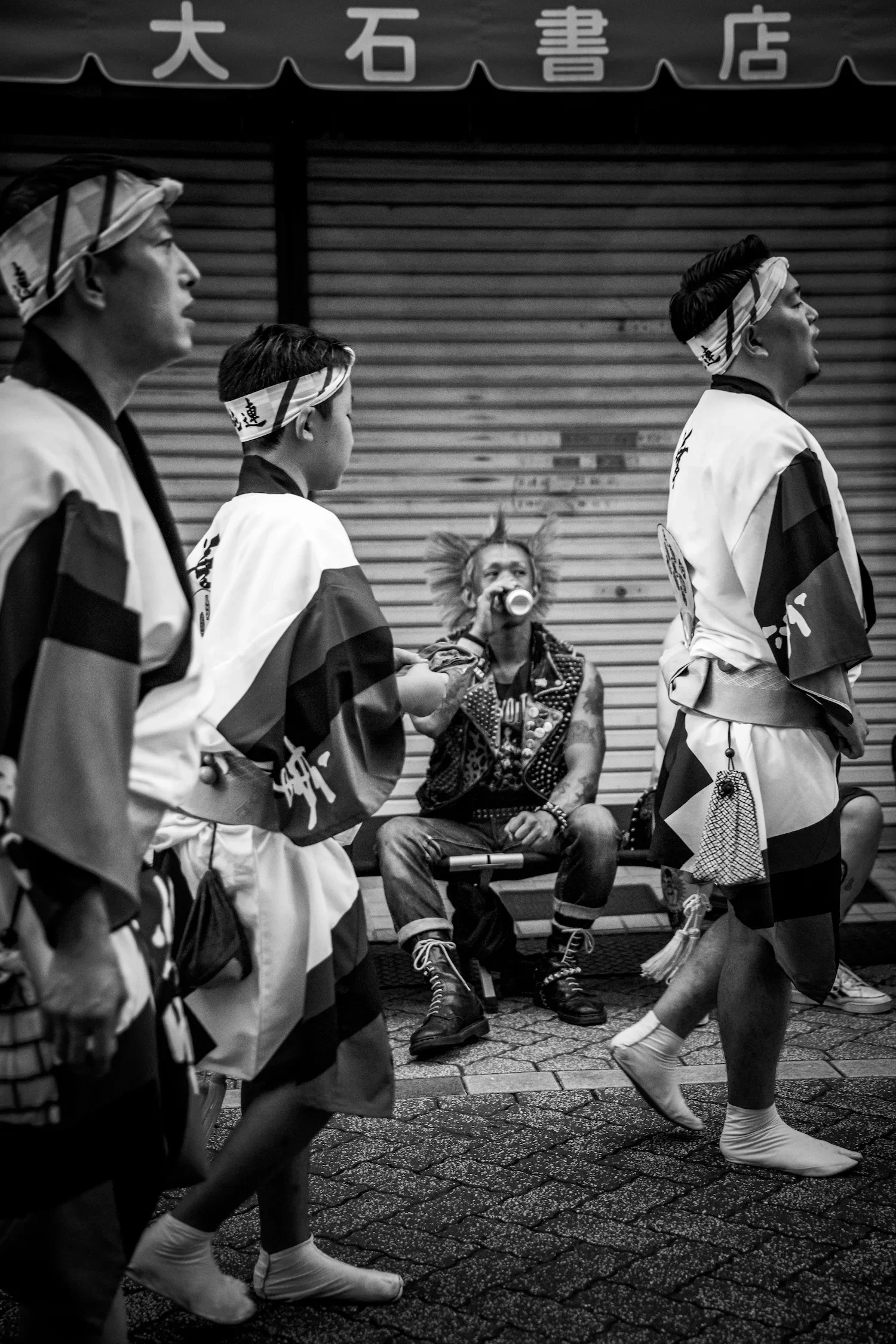 Black and white photo of three dancers in traditional Japanese costumes, walking in a line, with a woman with distinctive hair and makeup sitting on a bench drinking from a cup in the background. The scene appears to be in an indoor space with a closed shop with Japanese characters above.