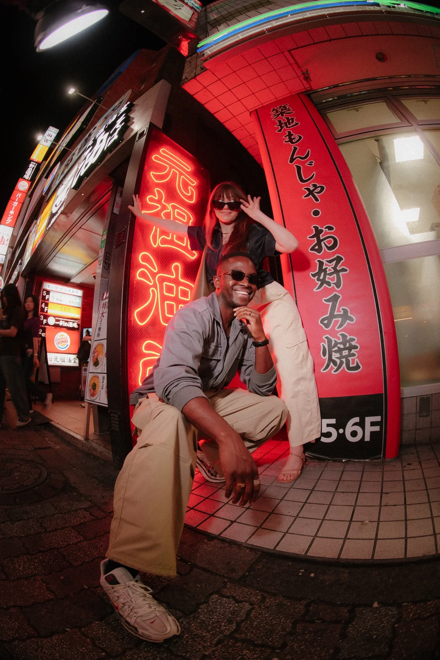 Two people, a woman and a man, are posing in front of a brightly lit red and black sign with Japanese writing outside a building at night. The woman is standing behind the man, wearing sunglasses, a black jacket, and beige pants, with her arm around his shoulder. The man is sitting on the ground, wearing sunglasses, a gray shirt, and beige pants, with a smile, resting his chin on his hand.