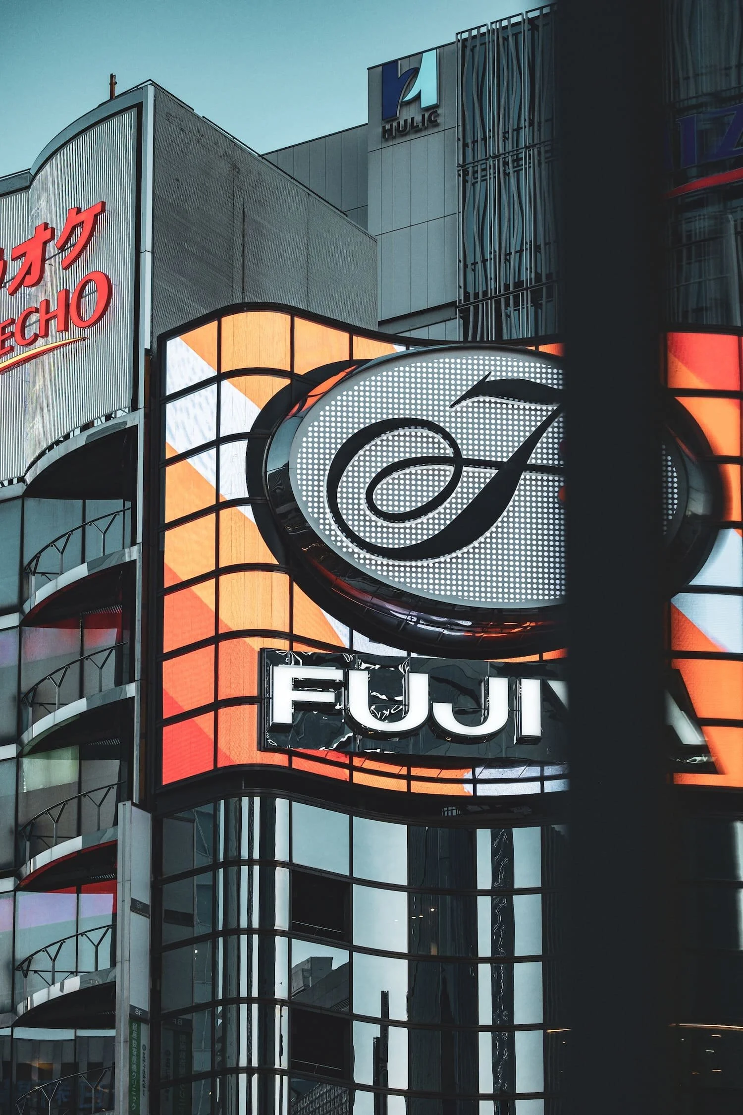 City building signage with illuminated signs reading 'Fujit', a large stylized letter 'A', and part of a sign reading 'ECHO' in Japanese characters. Modern glass and metal architecture reflect the surrounding cityscape.