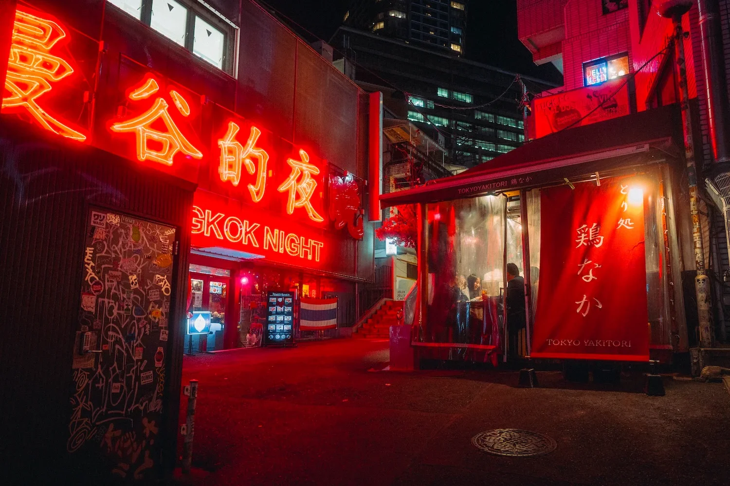 Nighttime street scene with neon red signs in Japanese and English, including 'SKOK NIGHT.' A small illuminated bus stop shelter with transparent walls has people inside. The area is dimly lit, with high-rise buildings visible in the background.