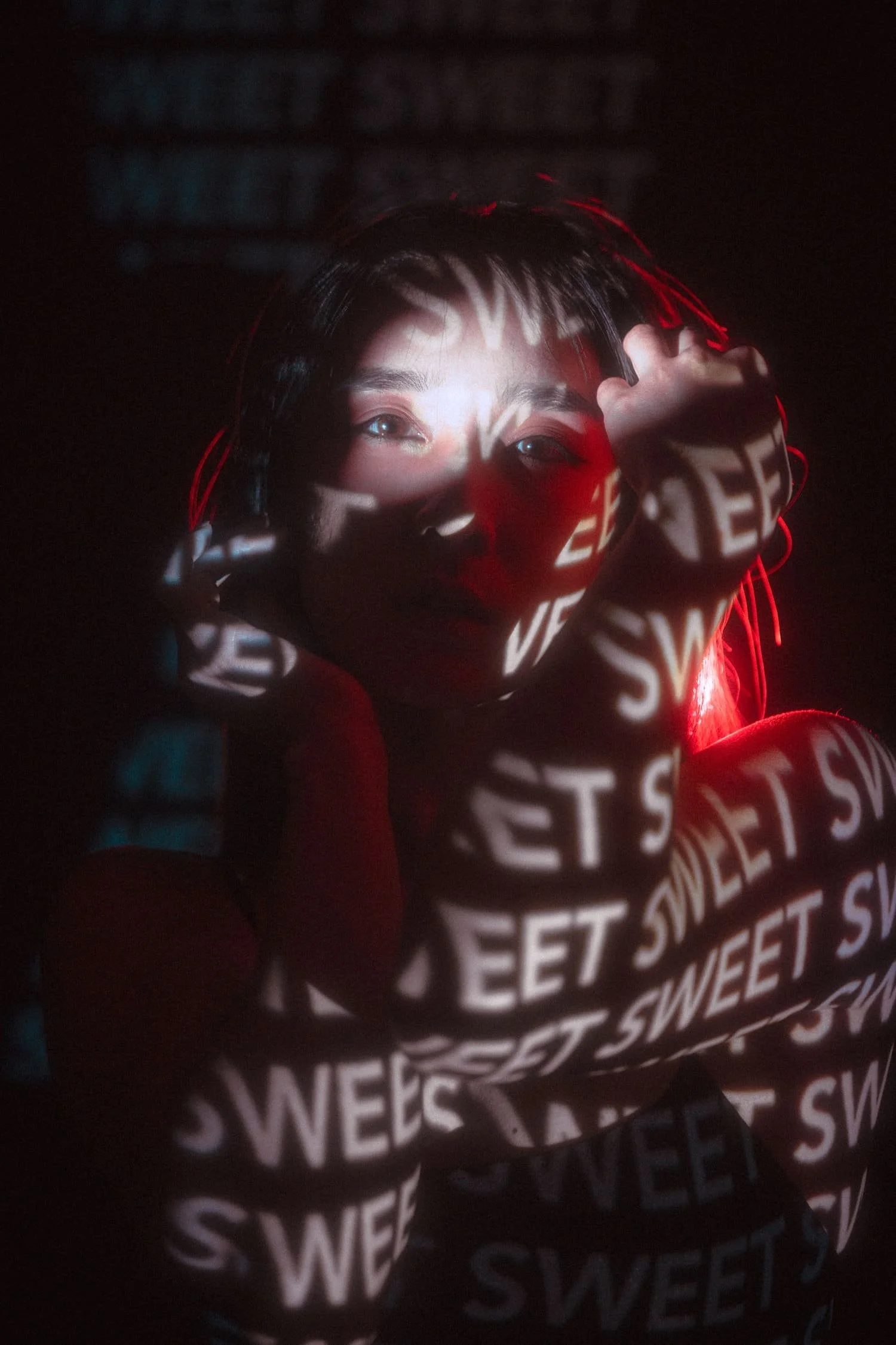 A woman with short dark hair faces the camera, with the words 'SWEET' projected onto her face and body in white letters. She has a calm expression and is holding her face with both hands, illuminated by red and white light effects.