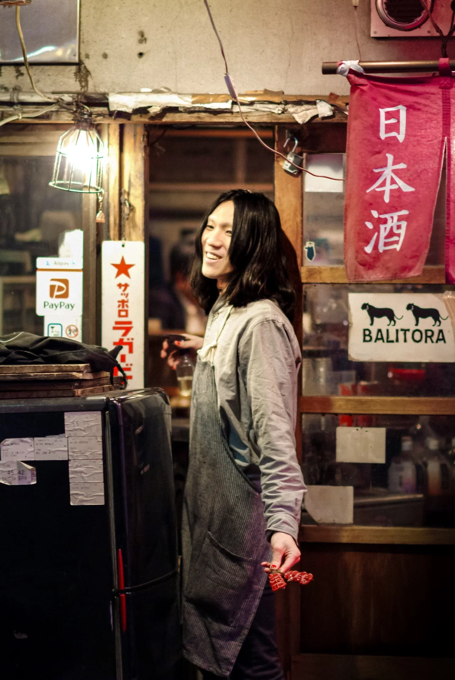 A woman with dark hair smiling while standing inside a restaurant or bar, with Japanese banners and signs on the wall behind her, some signs include logos for PayPay and Alipay.