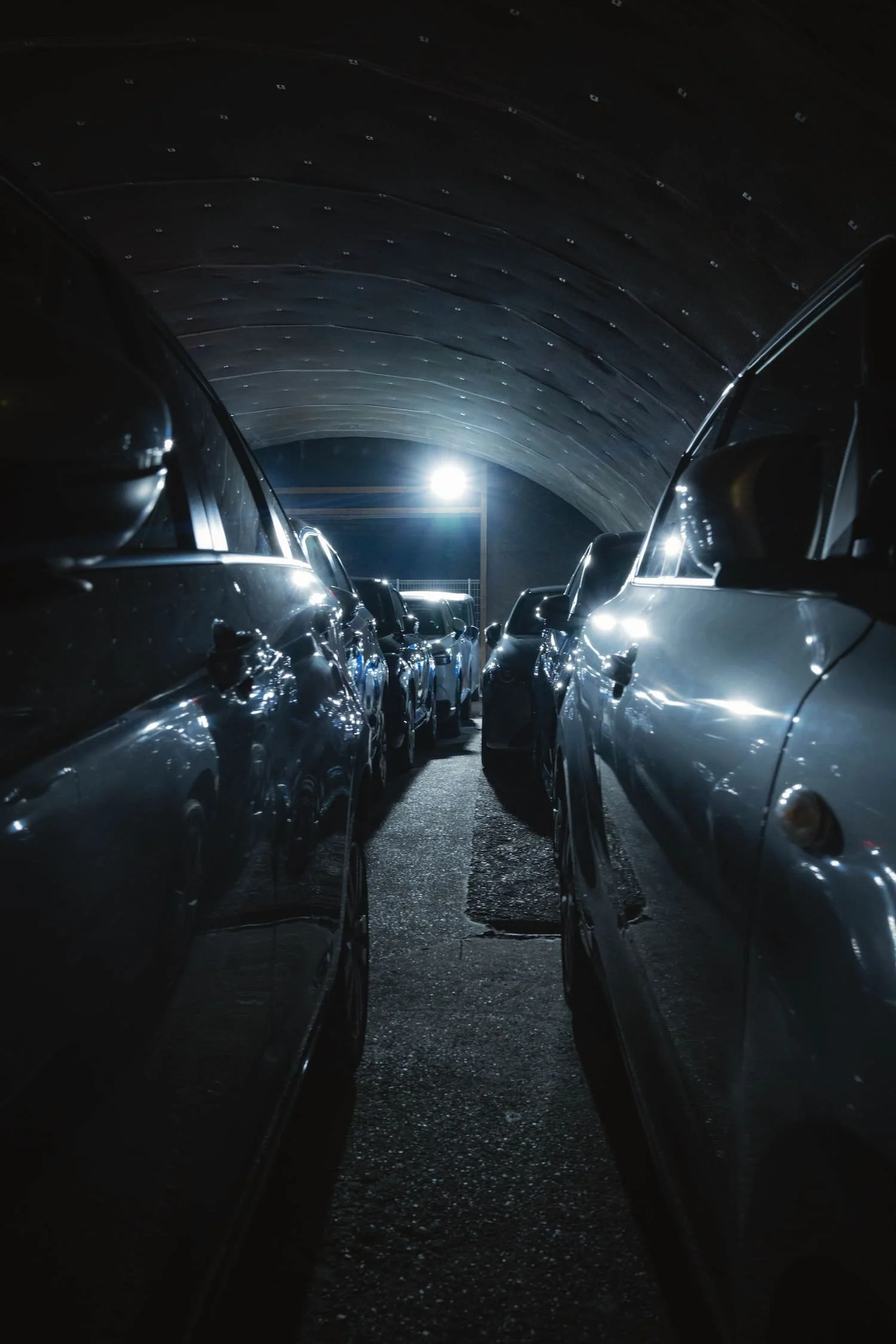 Dark underground parking garage with cars parked on either side, illuminated by a bright overhead light, arched ceiling visible. Tokyo street photography workshop