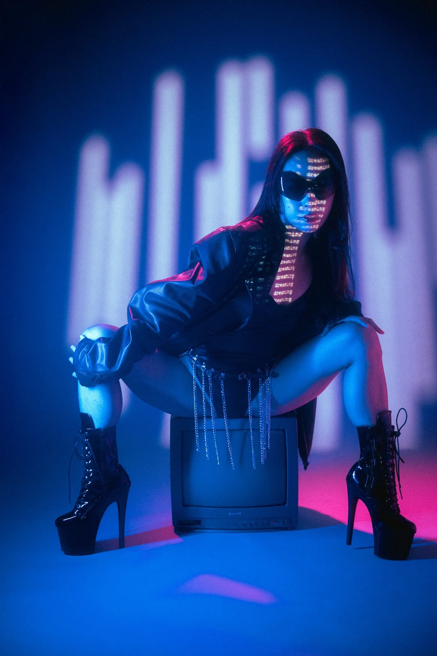 Woman with long dark hair wearing sunglasses, leather jacket, and high-heeled boots, squatting on a television set with neon lighting and an abstract background.