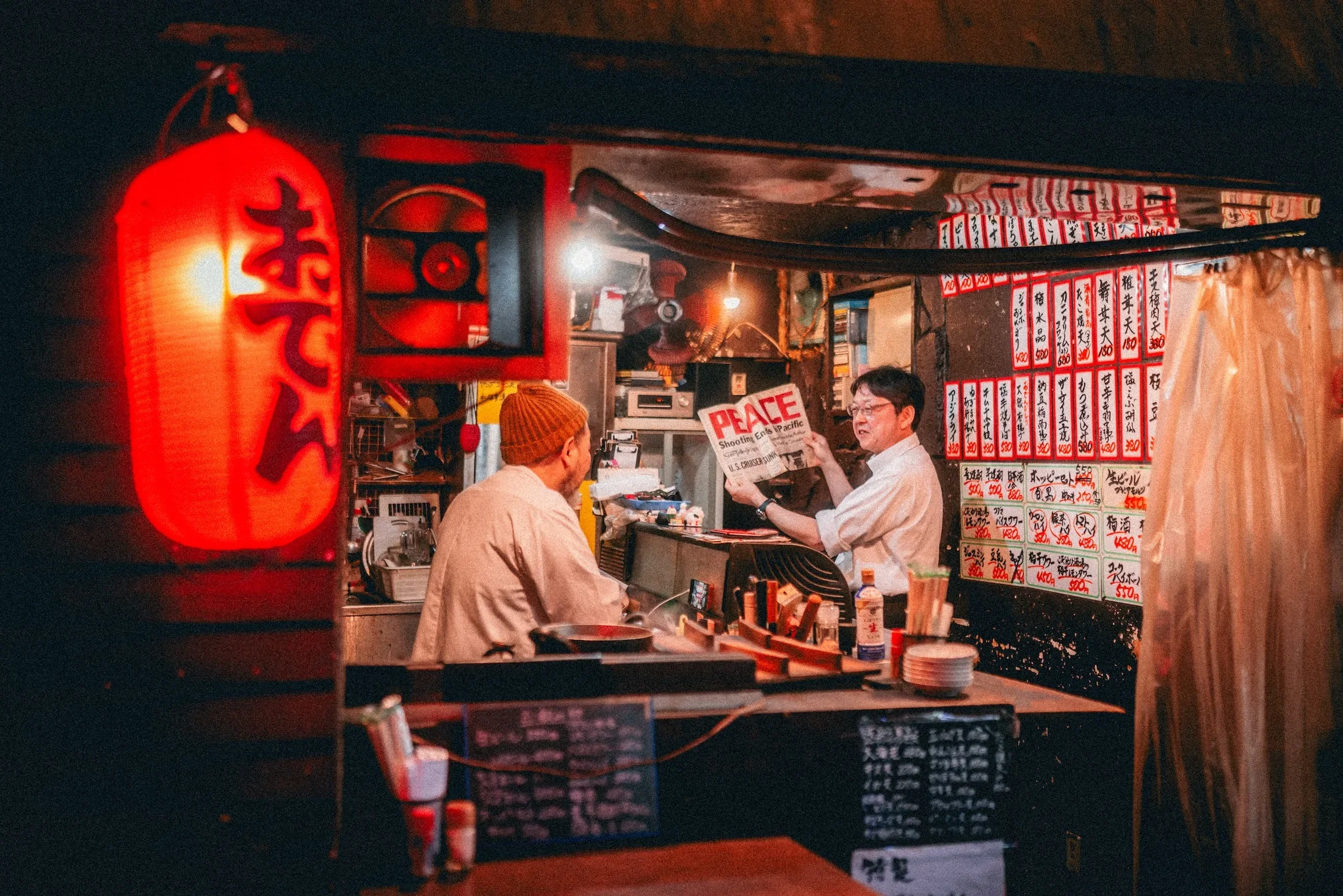 A Japanese restaurant with a red lantern, menu written in Japanese, and two men sitting at the counter, one reading a newspaper and the other speaking, inside a cozy, warmly lit space.