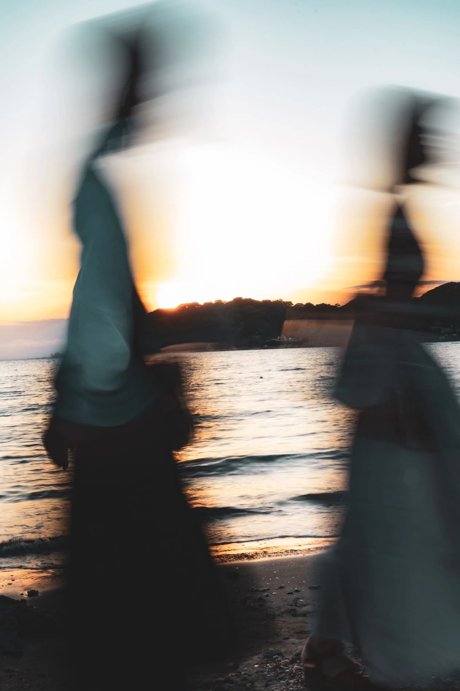 Two silhouetted people walking on the beach at sunset, with the ocean and a hillside in the background.