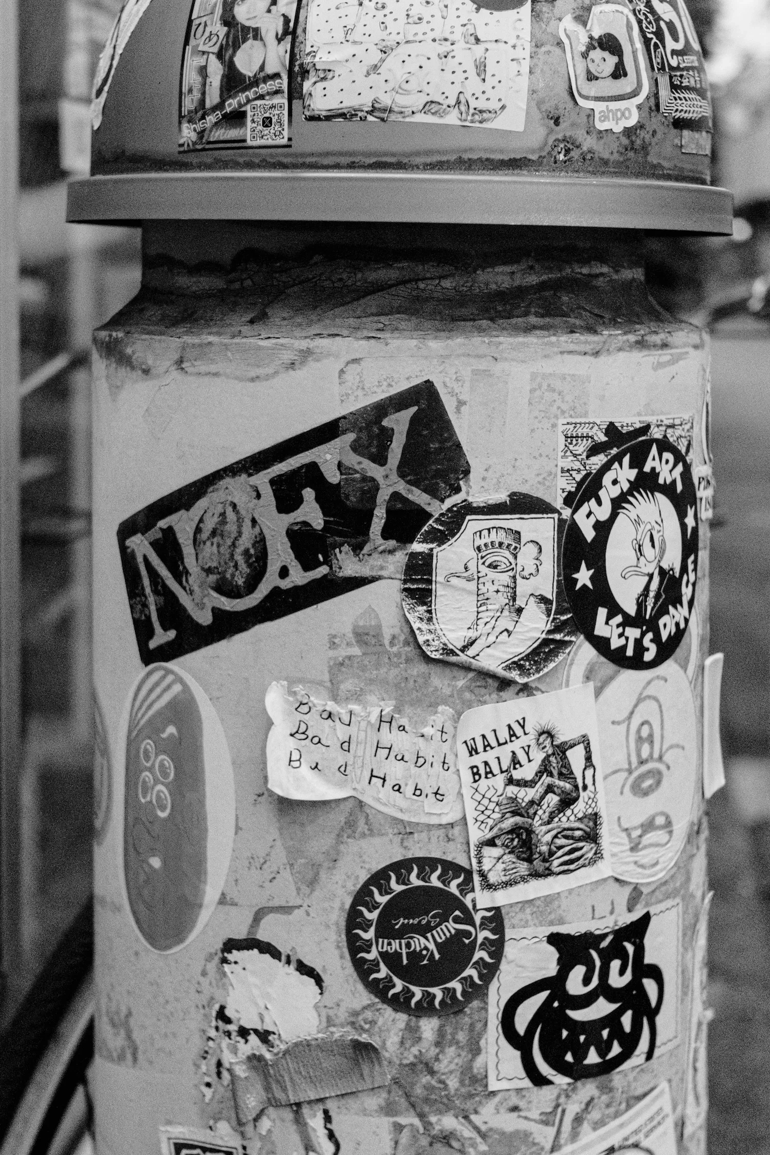 A collection of various stickers and torn pieces of paper are stuck on a surface, possibly a refrigerator, in black and white. The stickers feature different designs, including characters, logos, and handwritten notes.
