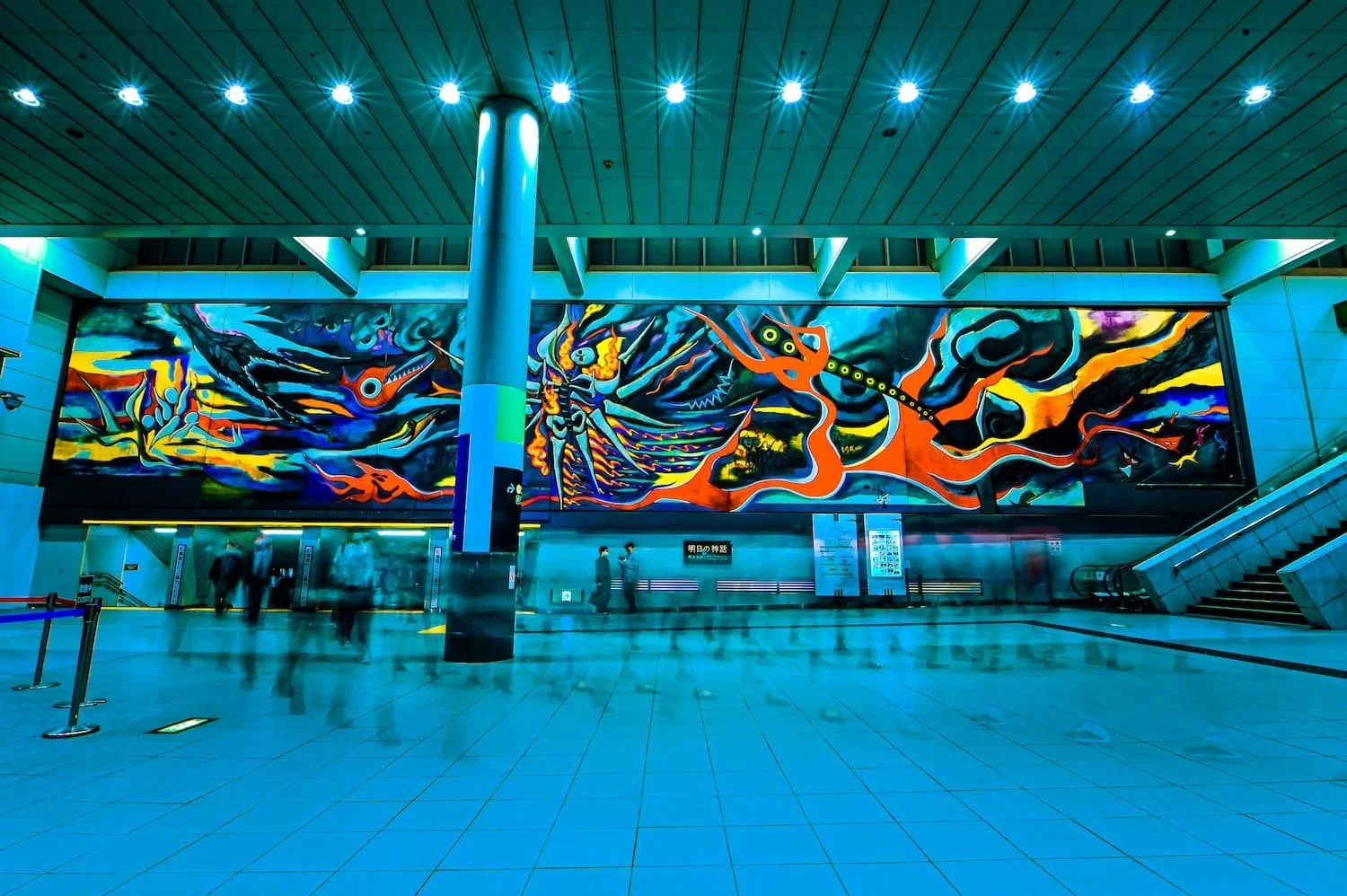 Colorful abstract mural inside a modern train station with people walking and an escalator on the right.