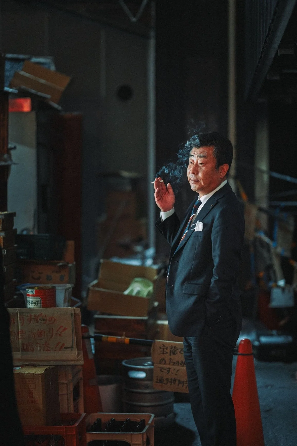A man in a suit smoking a cigarette in a dimly lit indoor setting, surrounded by boxes and equipment.