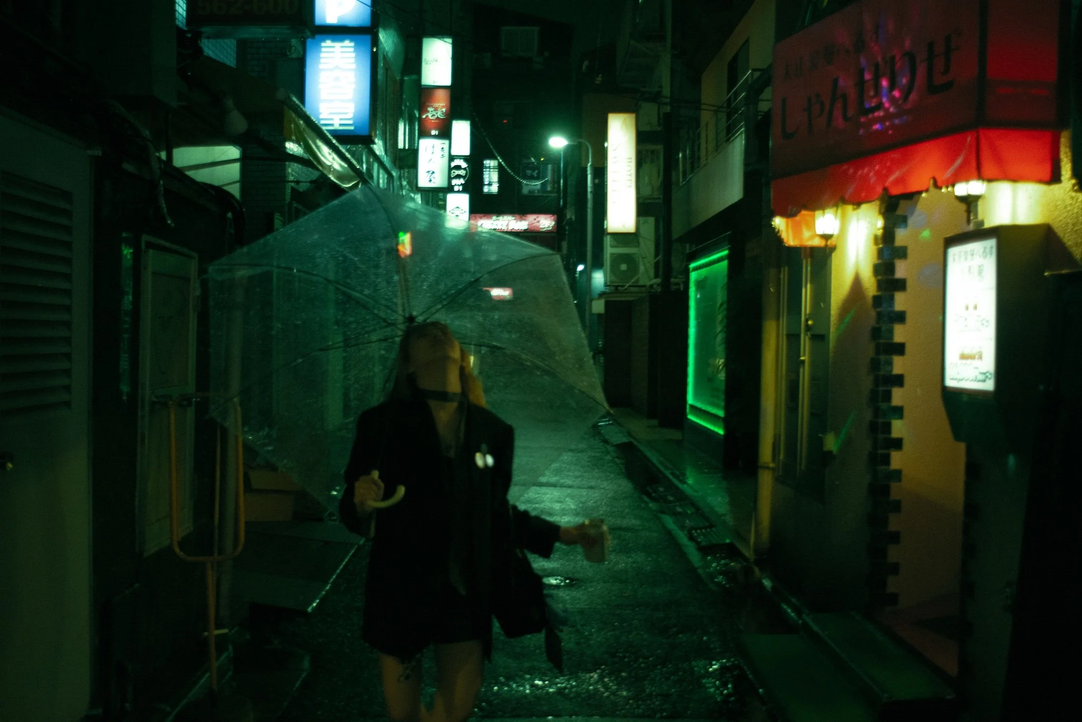 A woman holding an umbrella walking on a wet street at night surrounded by neon signs and shopfronts.