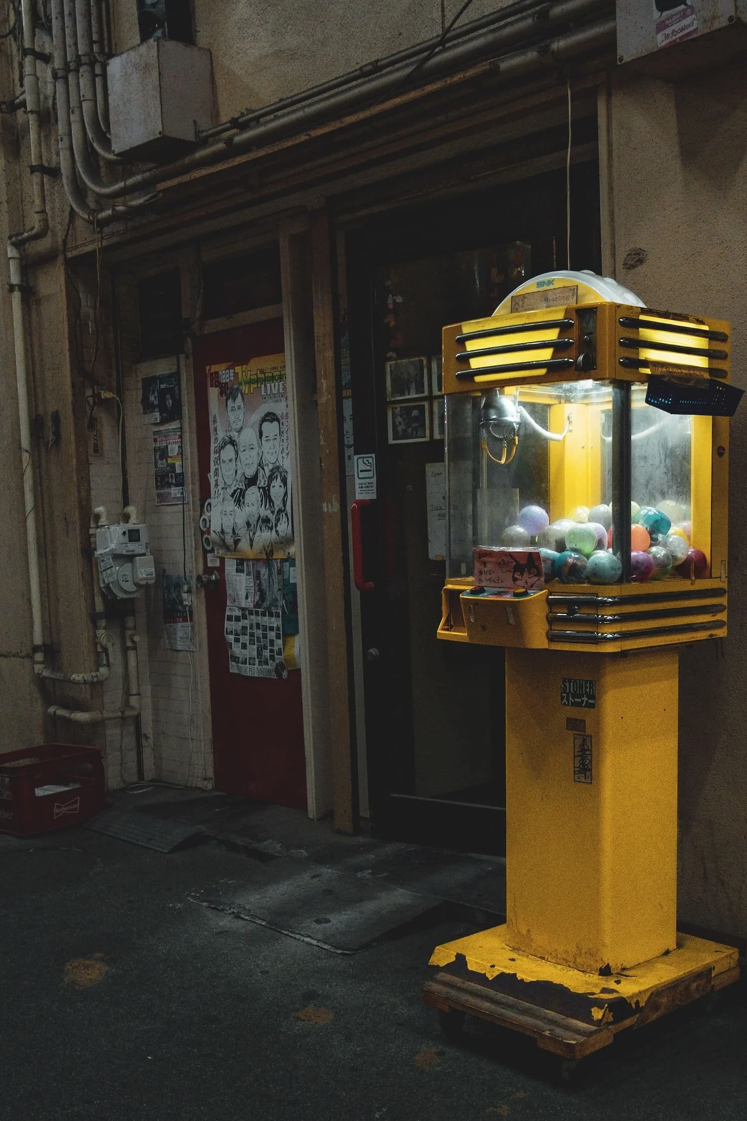 A yellow gumball machine filled with multicolored gumballs in front of a building entrance with posters and pipes.