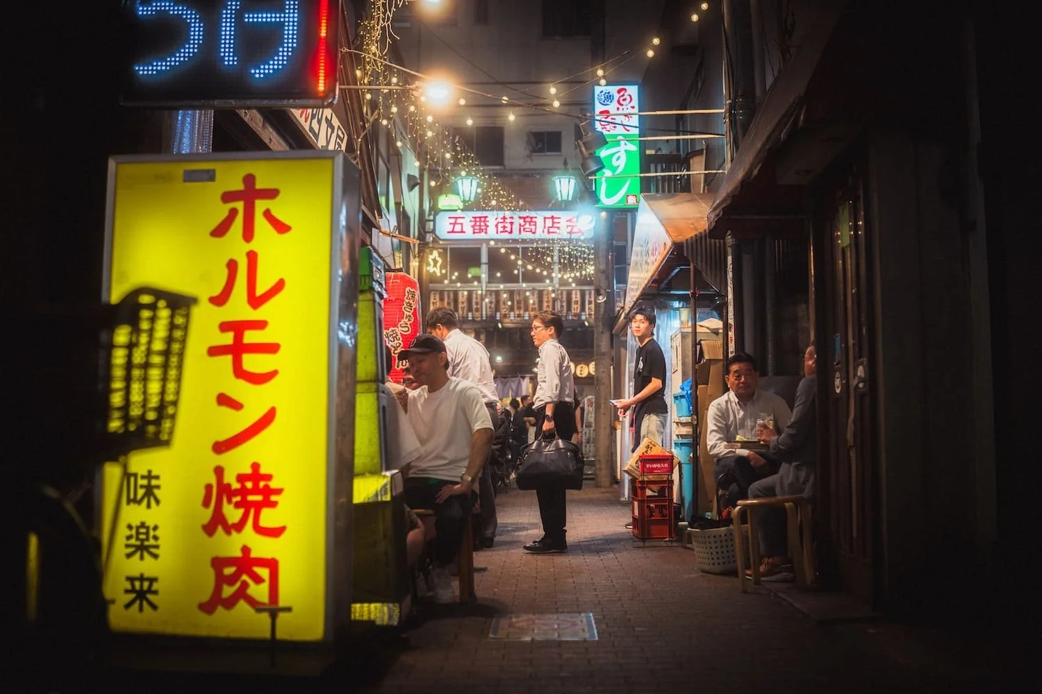 Night scene of a busy Japanese street with illuminated signs, people dining outside, and string lights overhead. Shot by Deniz Demir, Tokyo photographer.
