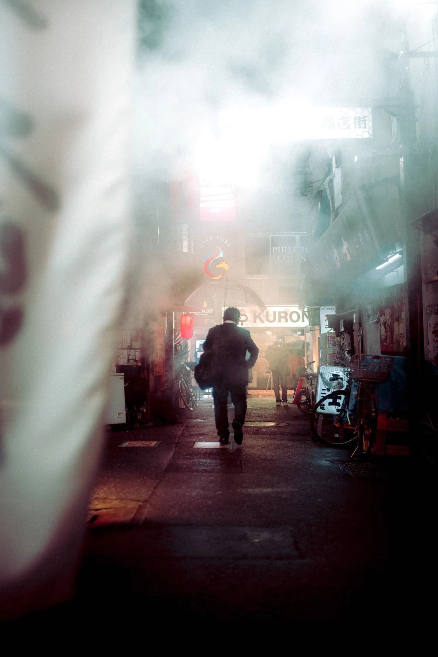 City street on a rainy night with a man walking, umbrellas, bicycles, neon signs, and fog.