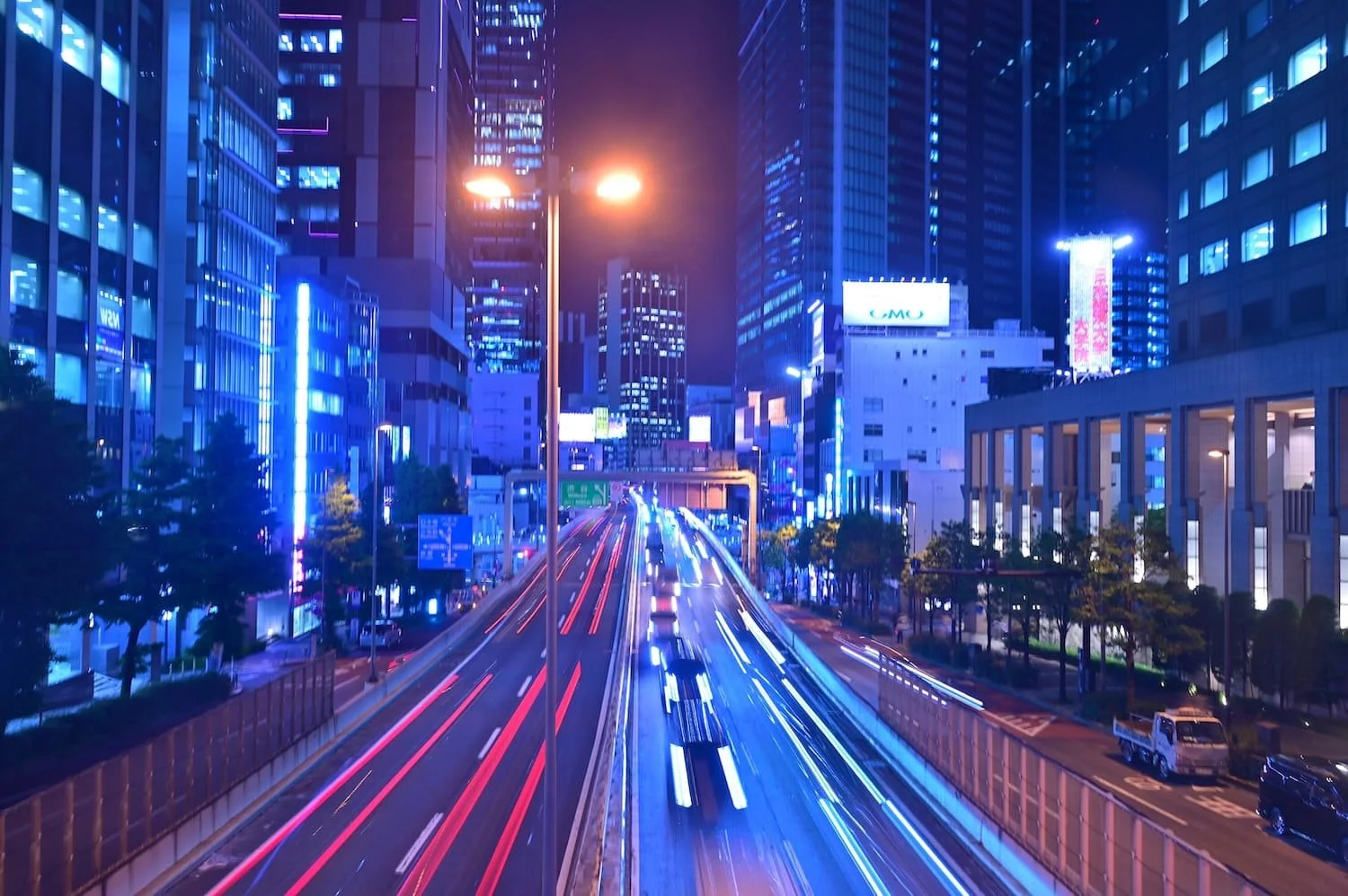 Nighttime cityscape with tall buildings illuminated by blue and purple lights, busy highway with streaks of red and white light from moving vehicles, and streetlights in the foreground.