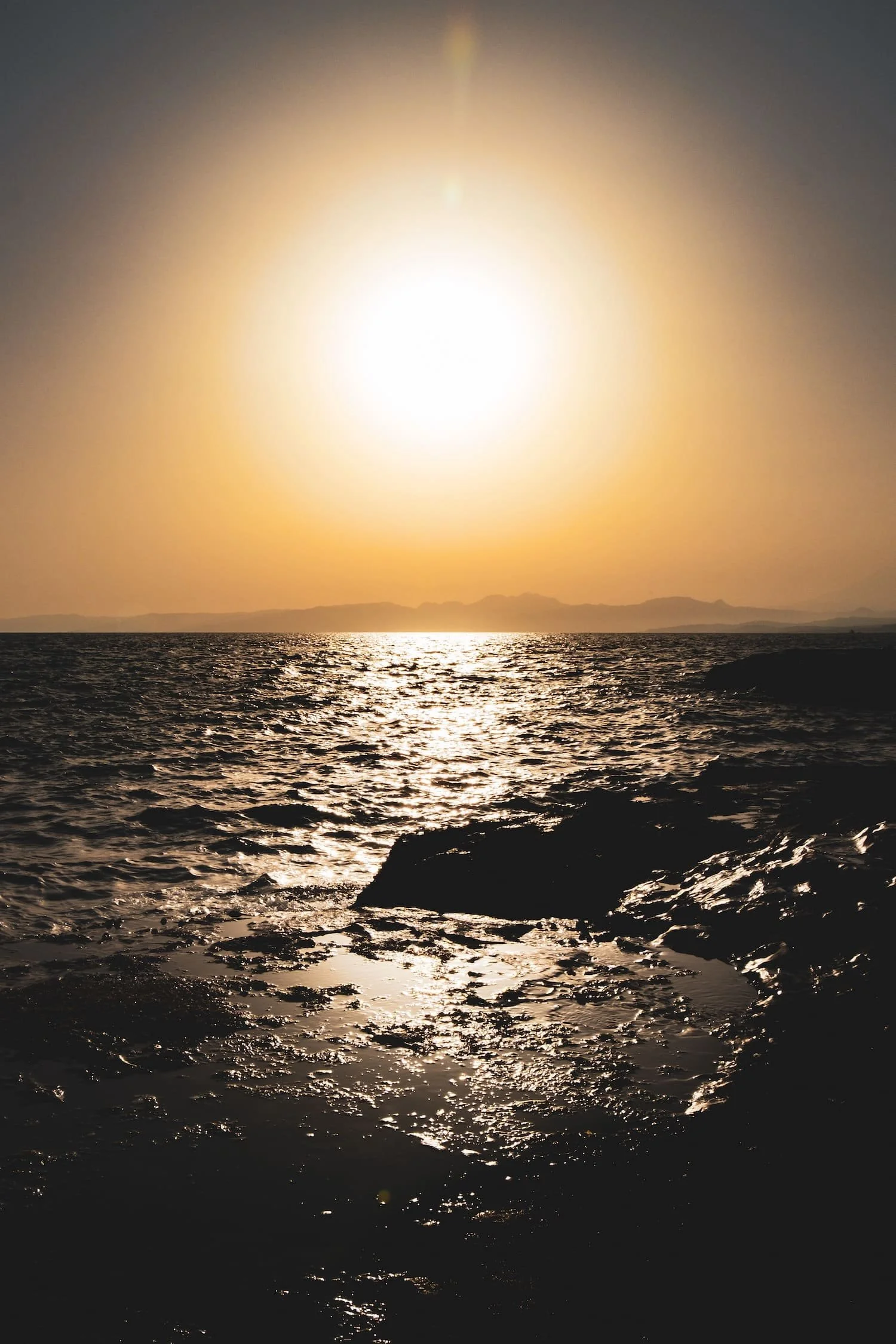Sunset over a rocky ocean shoreline with shimmering water and distant mountains in the background.