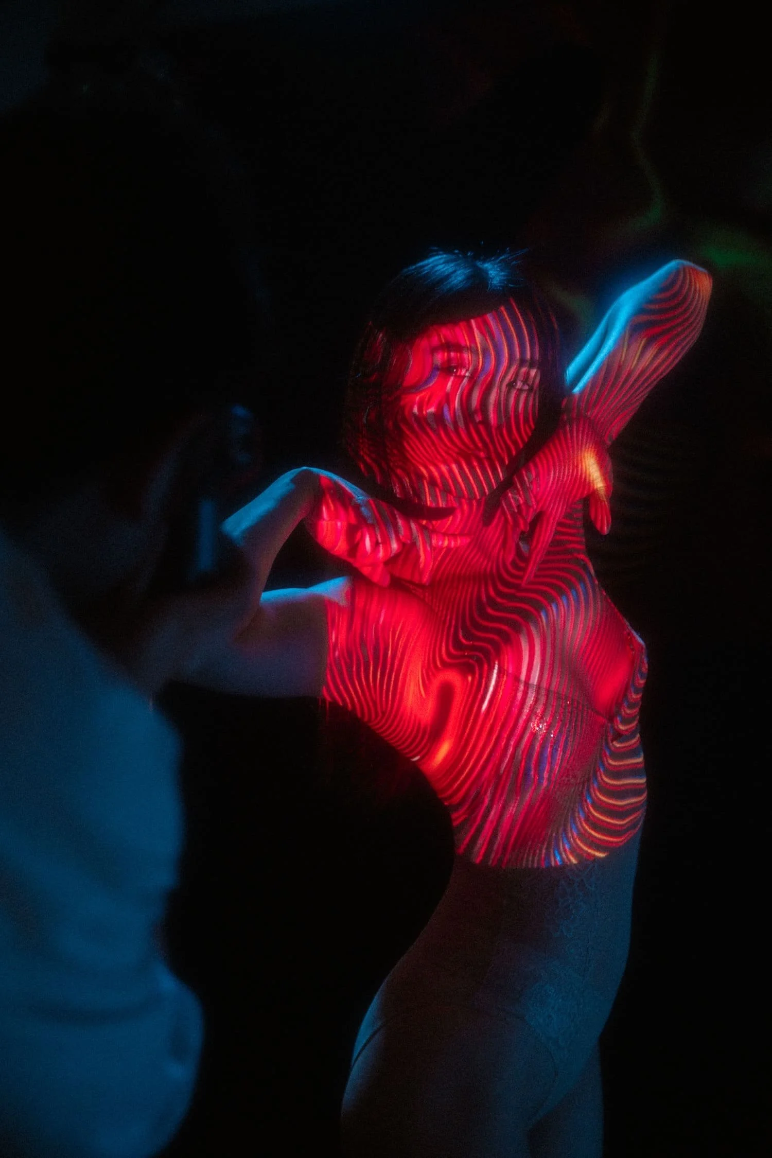 Behind the scenes shot of a a woman standing in a dark room with red and blue light projector images on her face and upper body, creating a vibrant, abstract pattern.