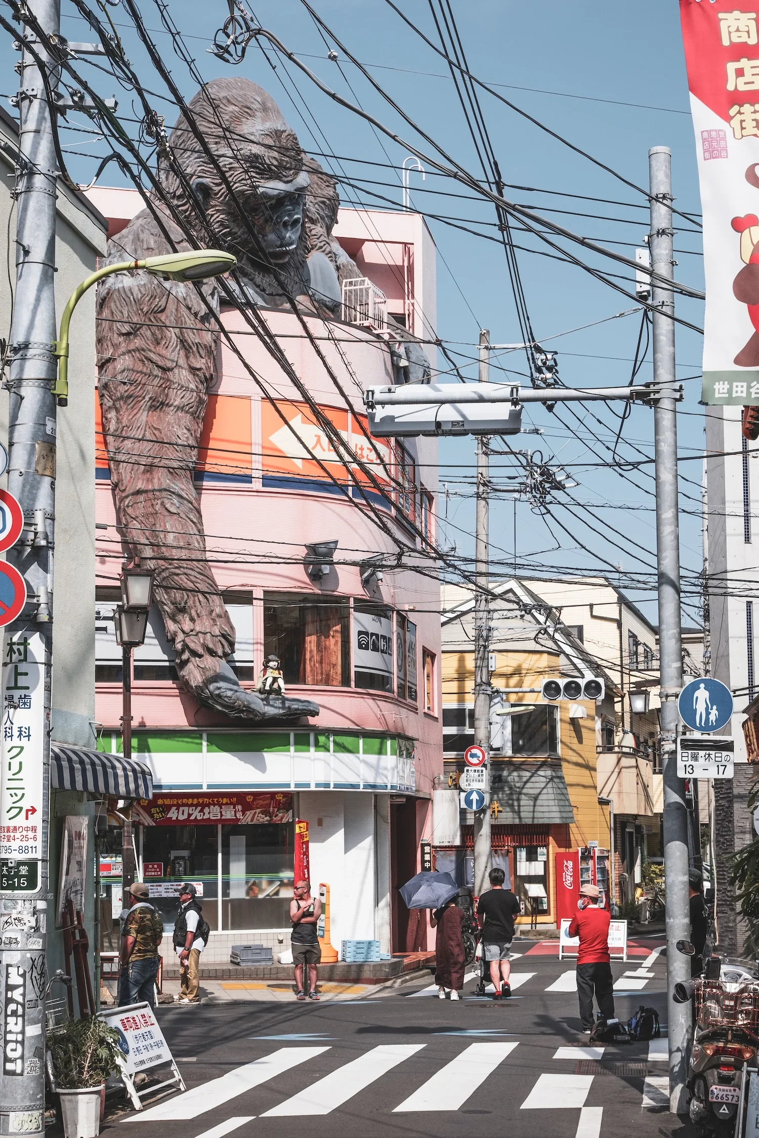 Street scene in Japan with pedestrians crossing a crosswalk, a building with a large gorilla statue on the upper floor, and various utility poles and signs.