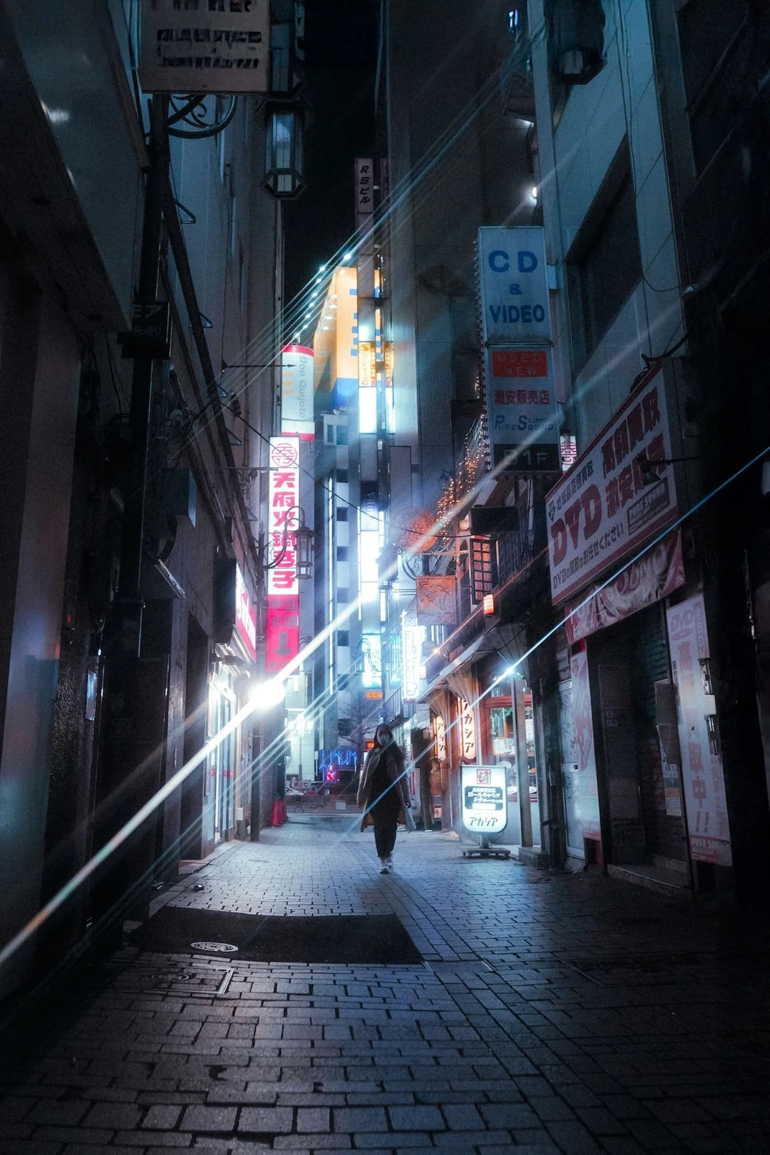 A person walking down a narrow, illuminated alley at night with colorful neon signs in Japanese and English on the buildings