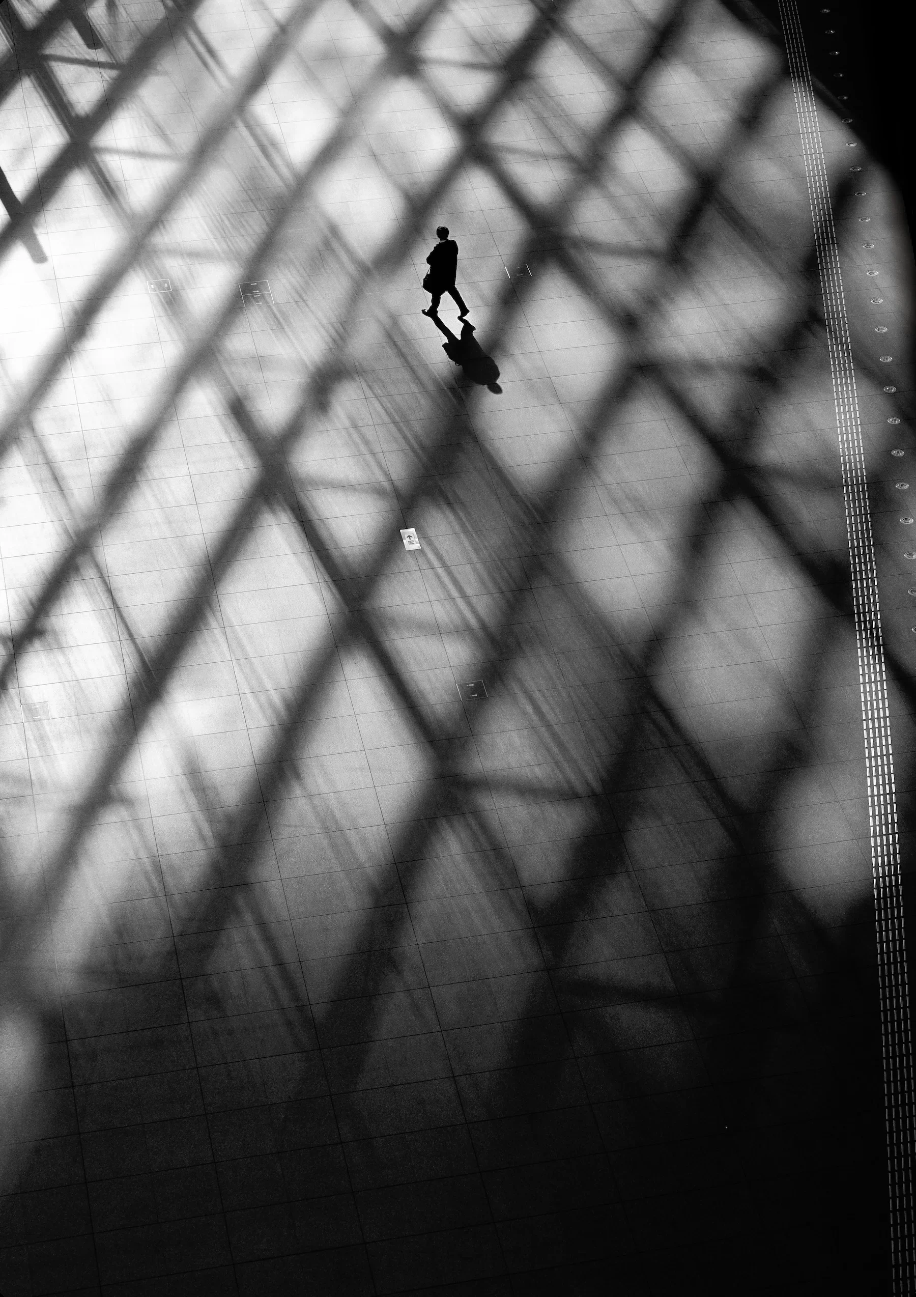 A person walking through an empty indoor space is captured from above through a fence or grille, creating a pattern of diagonal lines overlaying the scene.