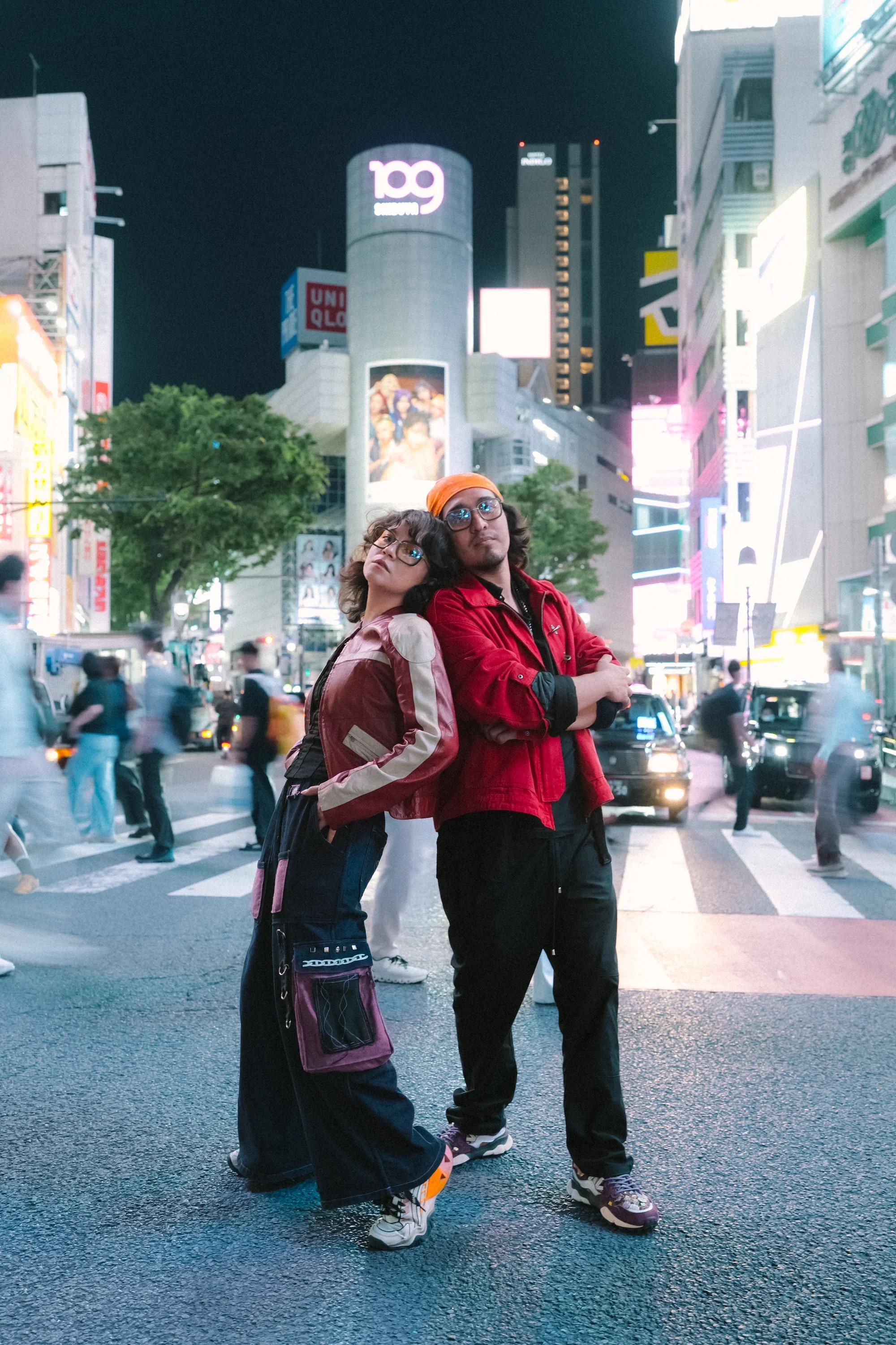 Two young people standing on a busy city street at night. The girl on the left has shoulder-length curly hair, glasses, a beige jacket with purple accents, and loose-fitting black pants. The boy on the right has shoulder-length wavy hair, glasses, a bright orange bandana, a red jacket, and dark pants. They are leaning their heads together, looking off into the distance. The background shows illuminated buildings, signs, and blurred pedestrians and vehicles.