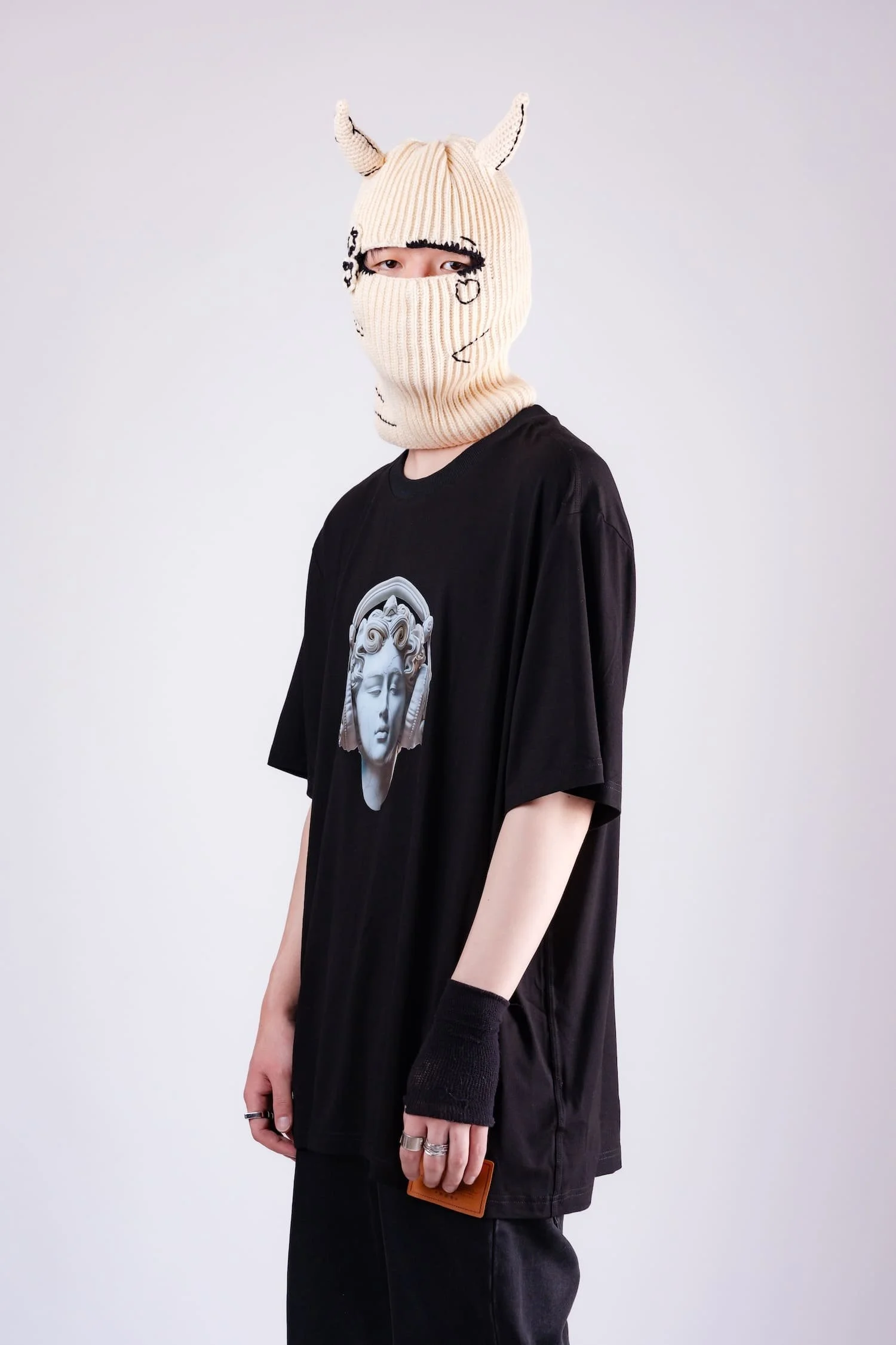 Studio portrait of a model wearing a beige horned knit balaclava mask and oversized black t-shirt featuring a classical statue graphic, photographed against a minimal white background by Tokyo photographer Deniz Demir.