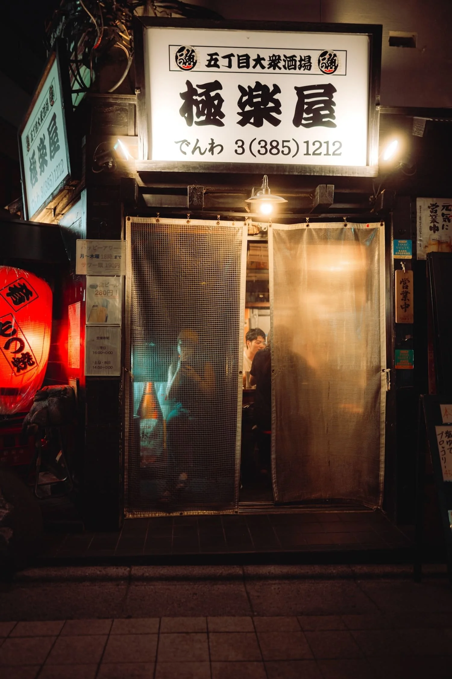 Nighttime view of a Japanese restaurant entrance with a bright sign displaying Japanese characters and a phone number, partially open noren curtains, and patrons visible inside.
