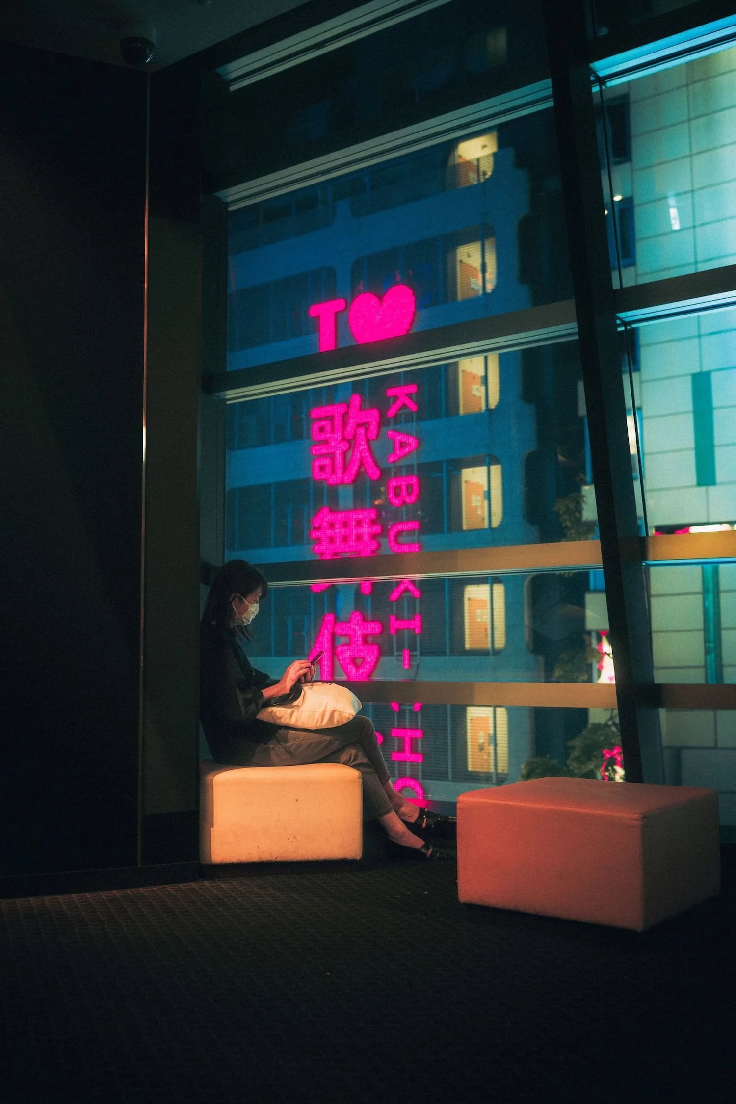 A woman sitting on a beige bench by large glass windows at night, using her phone. Neon pink and blue signs are reflected in the windows, including a hearts symbol and Japanese characters.