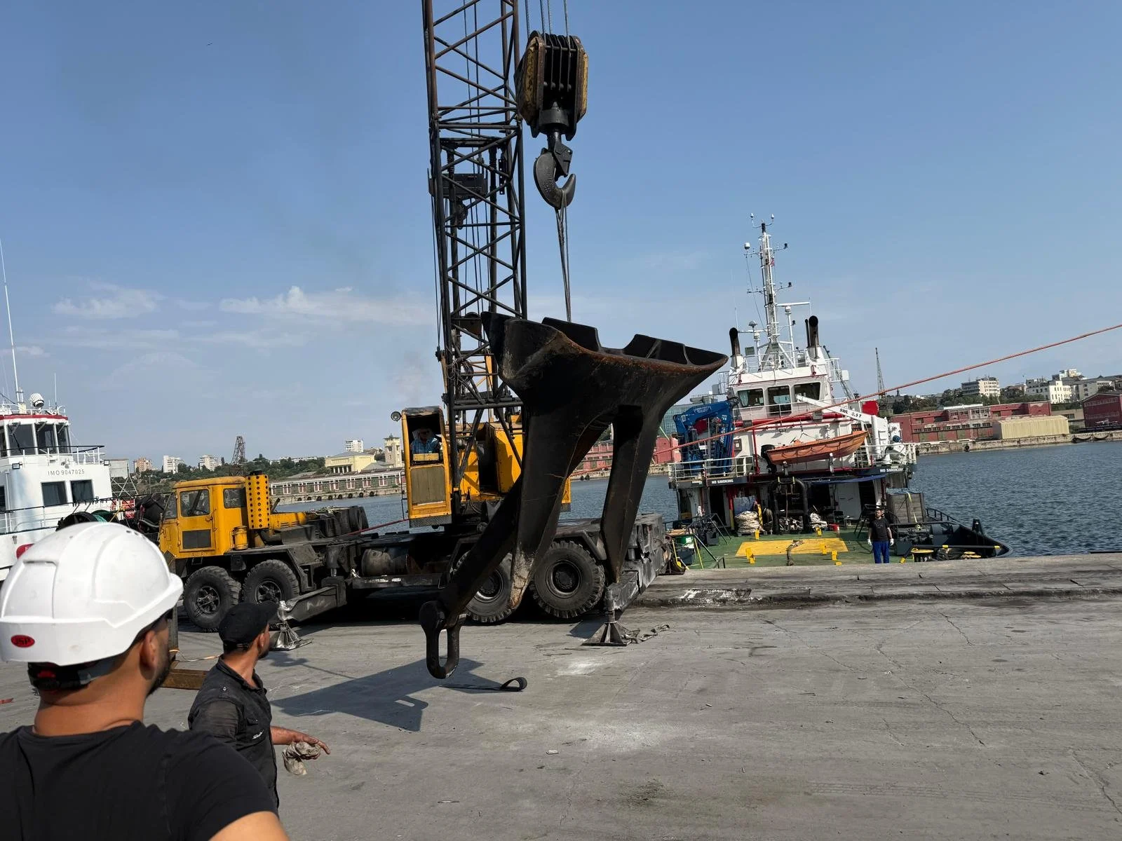 Construction workers observe a large anchor being moved by a crane at a dock with boats in the water and a cityscape in the background.