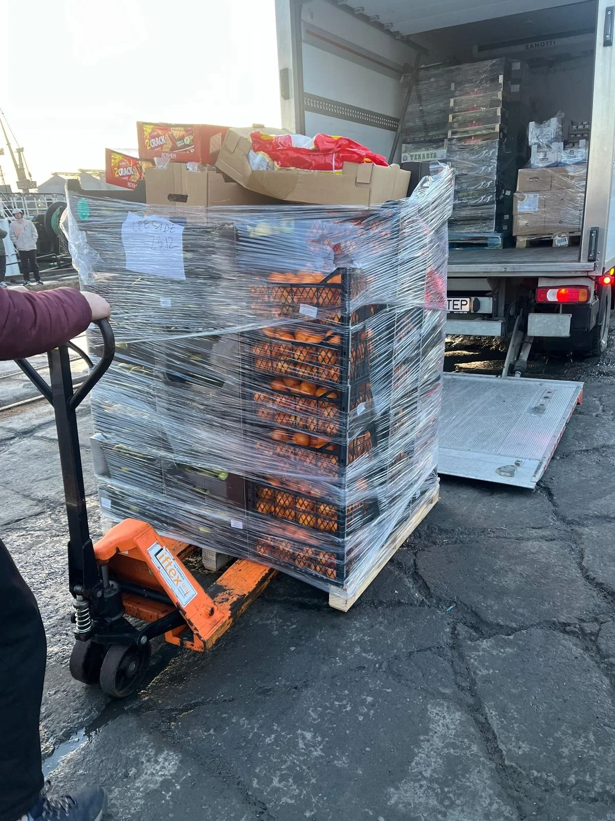 A pallet wrapped in plastic with crates of oranges, a box of snack crackers, and other items being loaded into a truck.