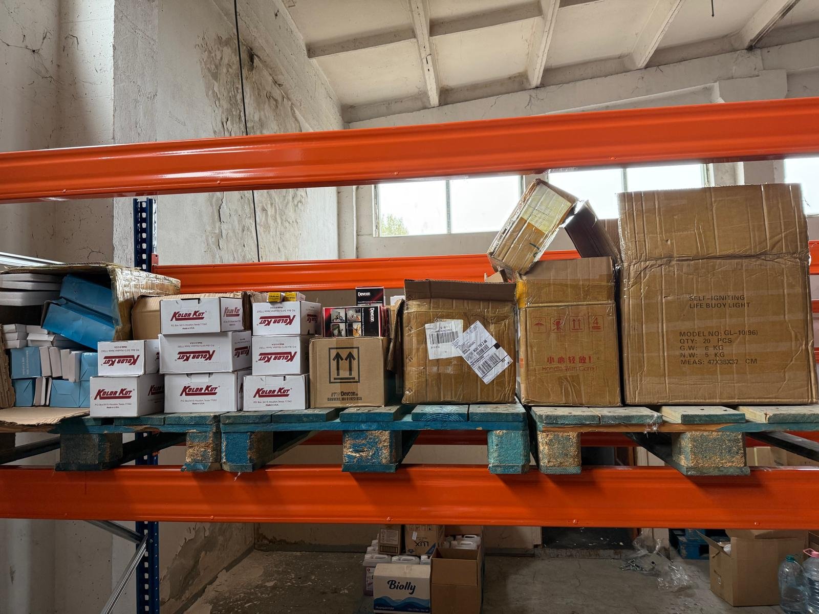 Warehouse shelf holding various boxes and packages, with orange metal supports, in an unfinished room with concrete walls and ceiling.