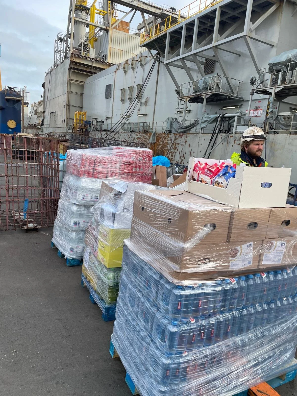 Pallets of bottled water and supplies on a dock next to a large, rusty ship, with a worker wearing a safety helmet and high-visibility jacket.