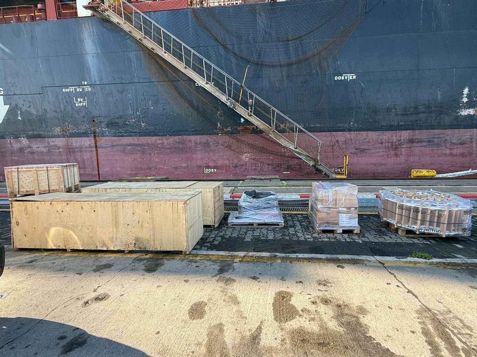 Bottles, wooden crates, and pallets of supplies on a dock next to a large ship with a red and black hull, and a metal gangway leading onto the ship.